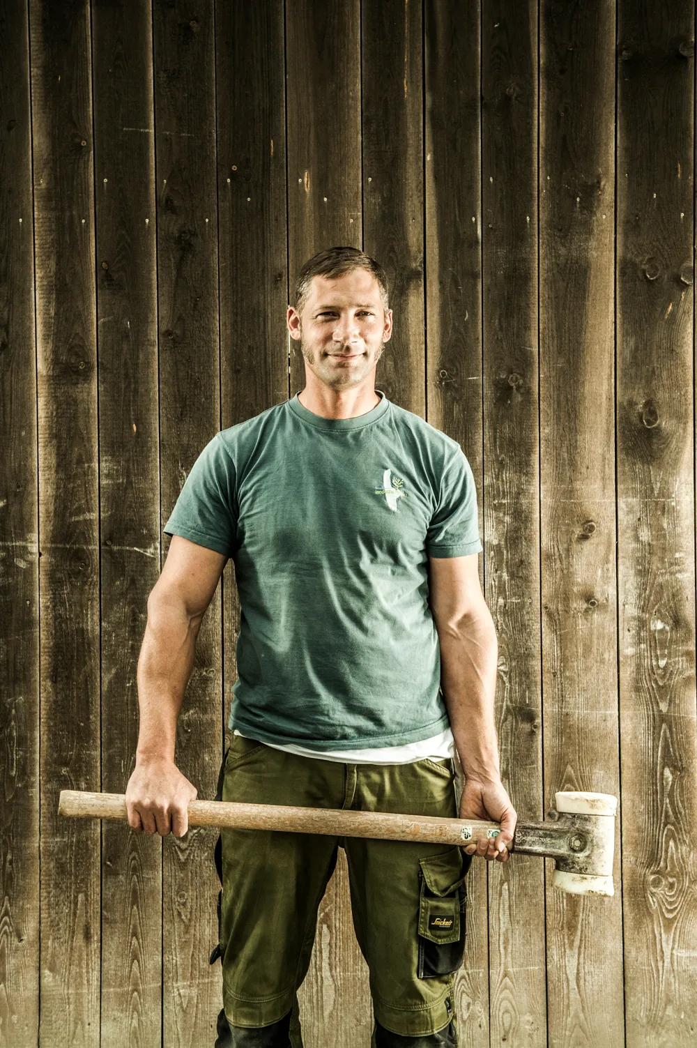 Team member in a blue T-shirt holding a sledgehammer in front of a weathered wooden wall — commercial and brand photography by Janina Laszlo for Ammerseer Landschaftsbau, landscaping company in Bavaria, Germany