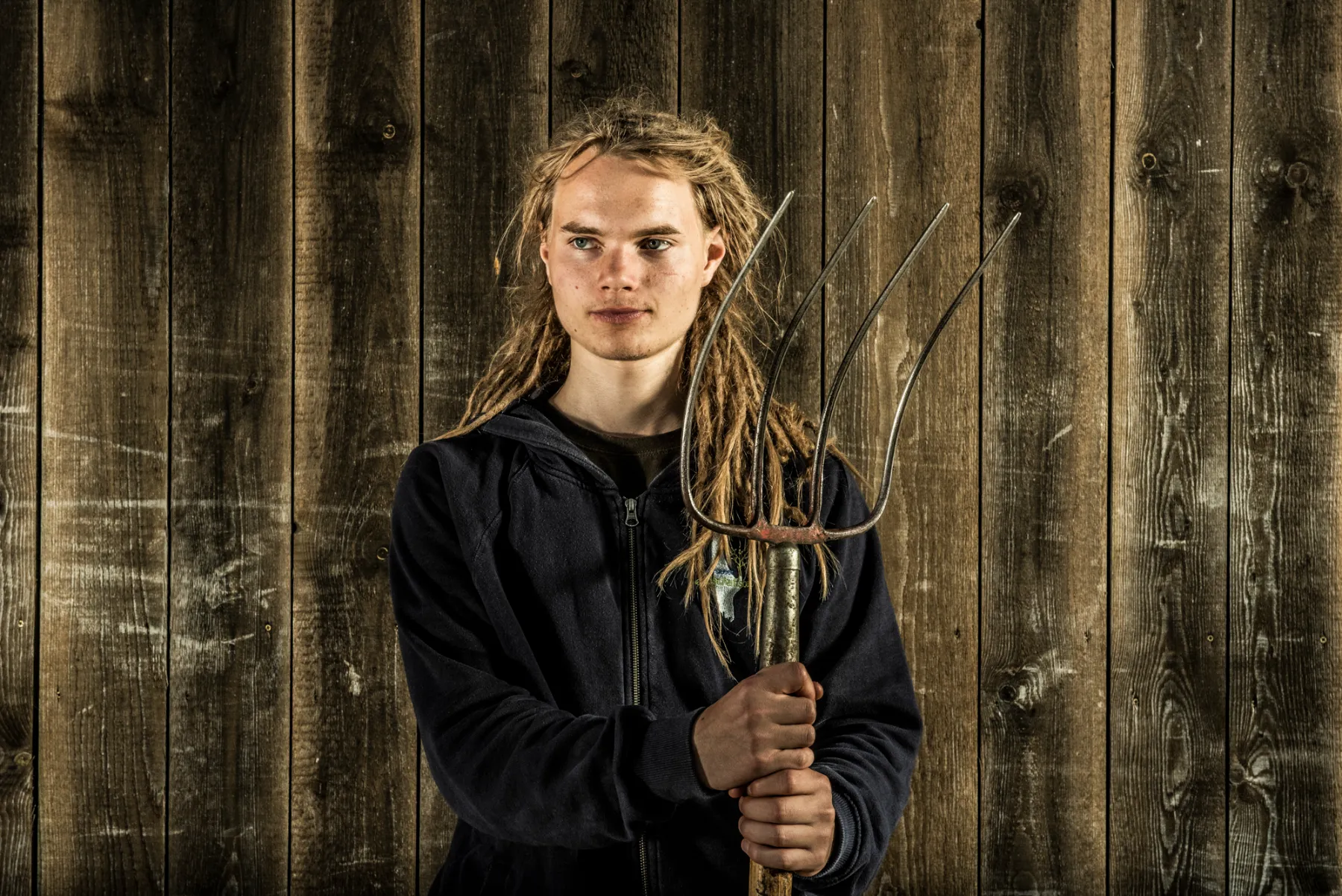Young team member with dreadlocks holding a pair of branches in front of a wooden wall — commercial portrait by Janina Laszlo for Ammerseer Landschaftsbau, Bavaria, Germany