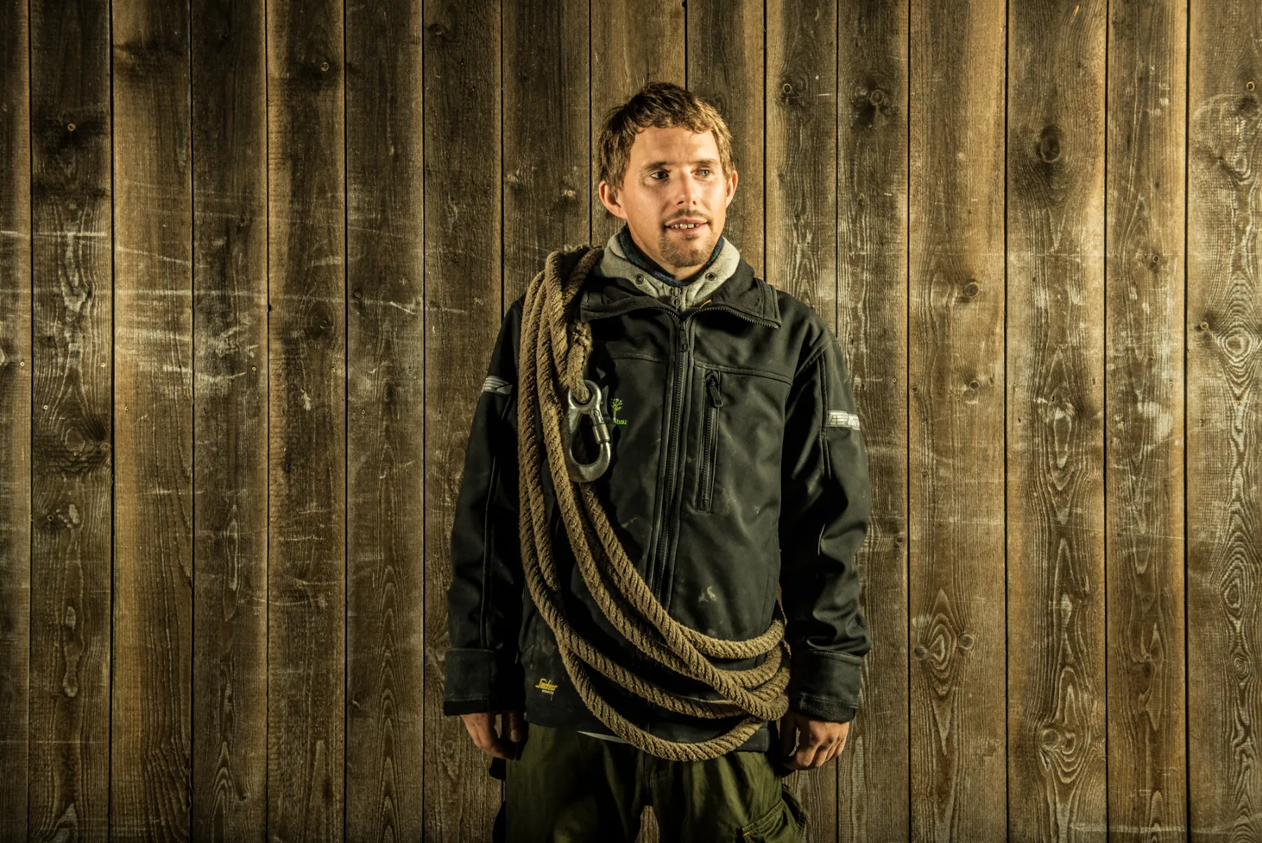 Team member with a coiled rope over his shoulder in front of a wooden wall — commercial portrait by Janina Laszlo for Ammerseer Landschaftsbau, Ammersee region, Bavaria