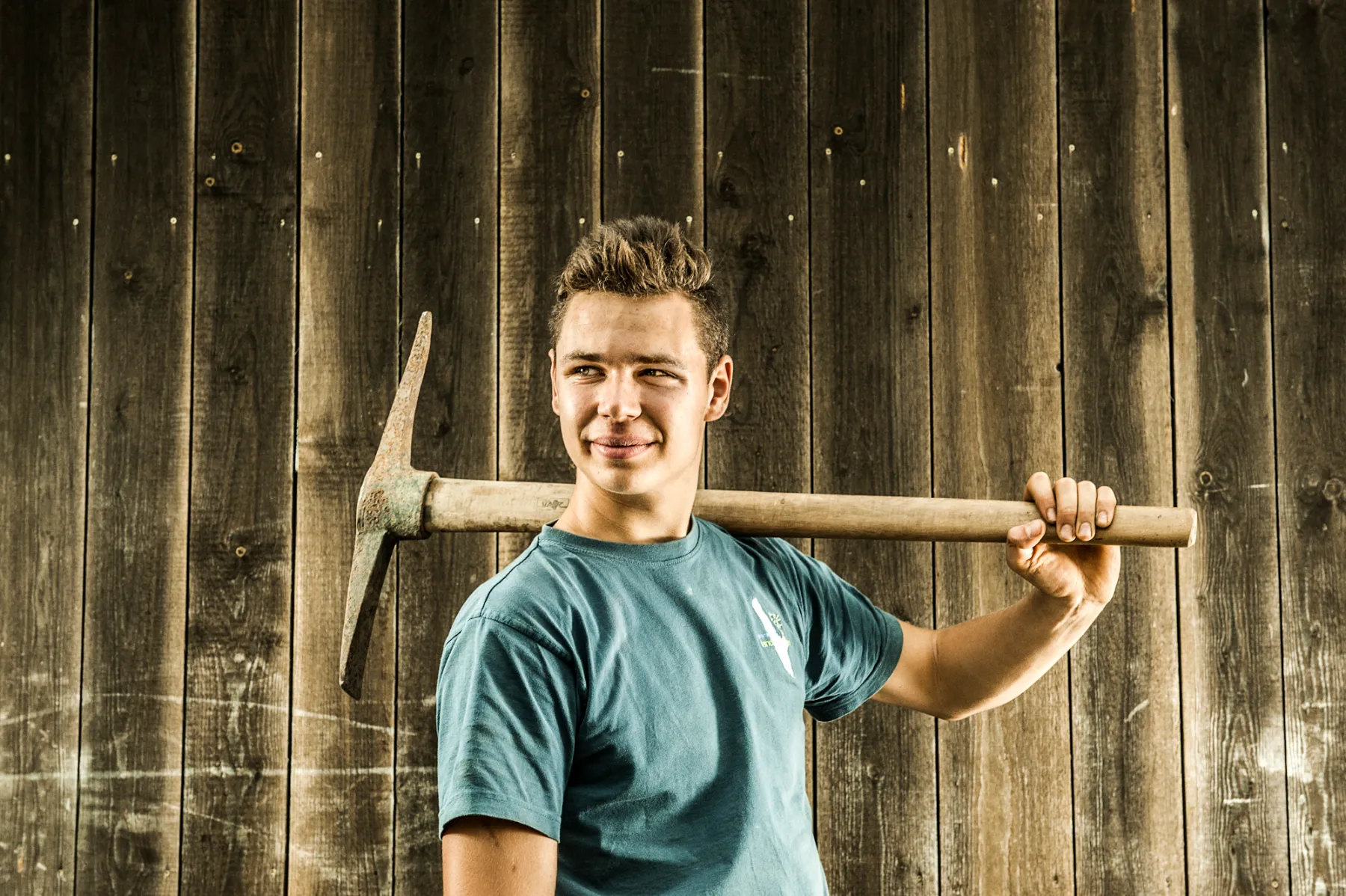 Young team member in a blue T-shirt with a pickaxe over his shoulders — brand portrait by Janina Laszlo for Ammerseer Landschaftsbau, Ammersee region, Bavaria