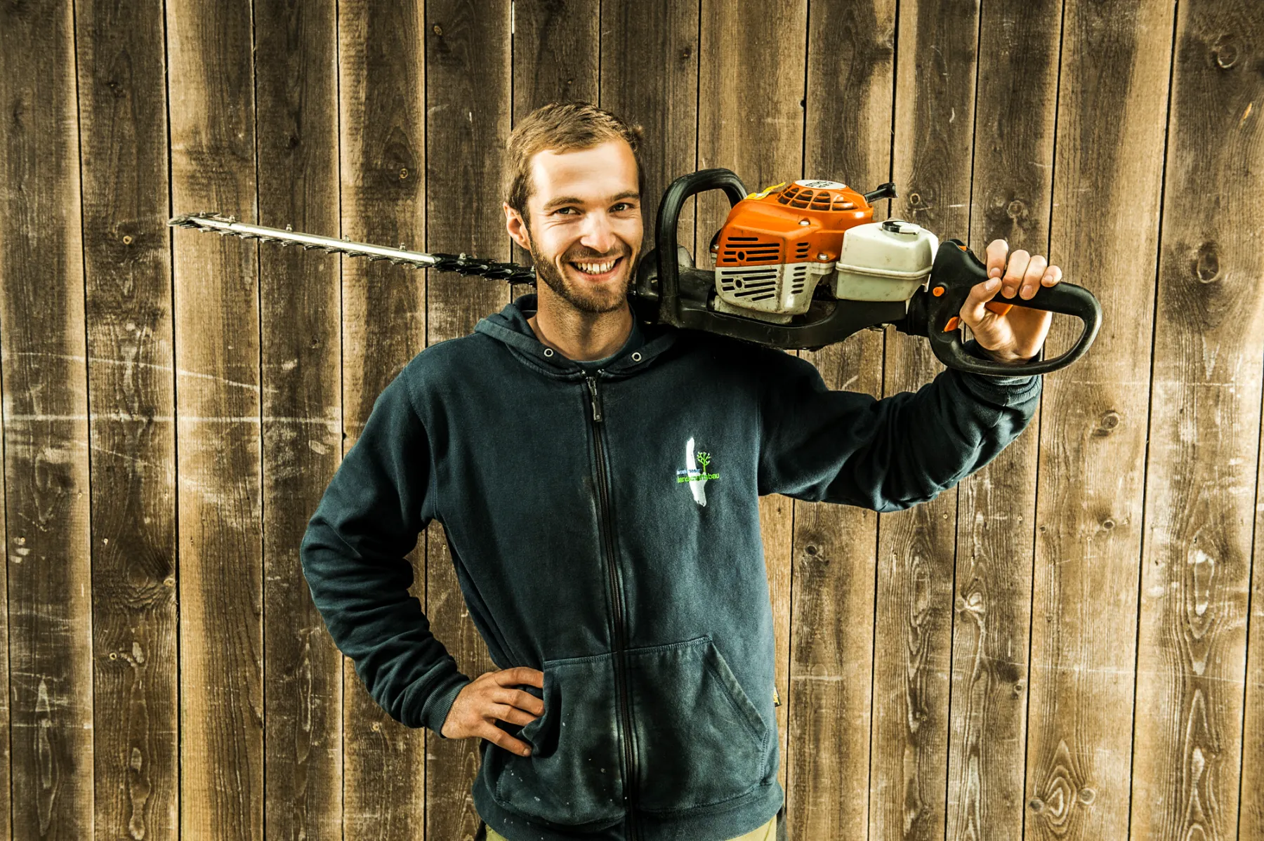Bearded team member in a navy sweater carrying a hedge trimmer — commercial photography by Janina Laszlo for Ammerseer Landschaftsbau, landscaping company in Bavaria, Germany