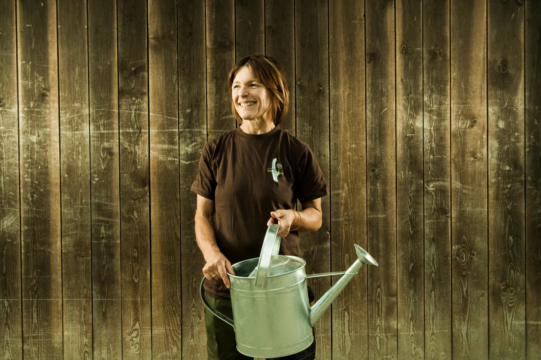 Woman with a metal watering can in front of a weathered wooden wall — brand photography by Janina Laszlo for Ammerseer Landschaftsbau, Bavaria, Germany