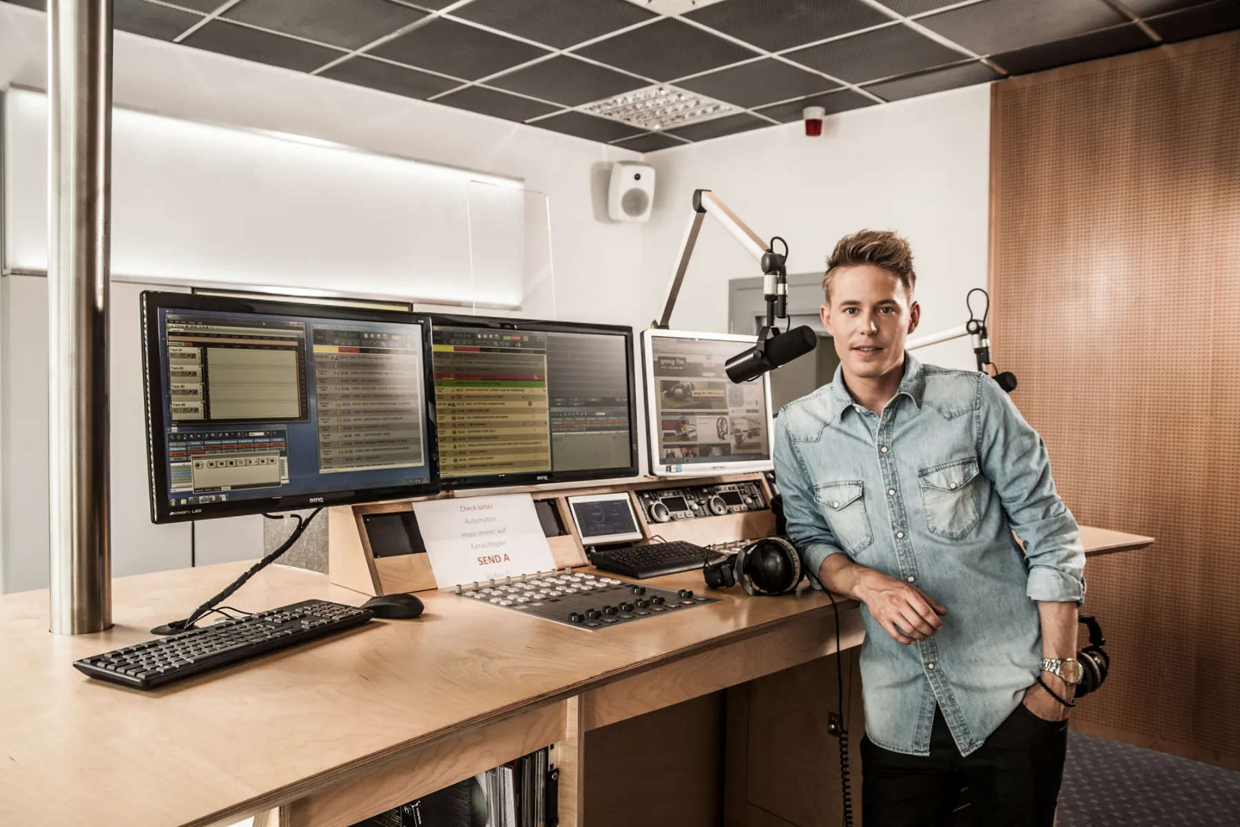 Timo Killer leaning on a radio studio desk beside monitors and microphones — editorial portrait for the AOK healthcare campaign by German photographer Janina Laszlo
