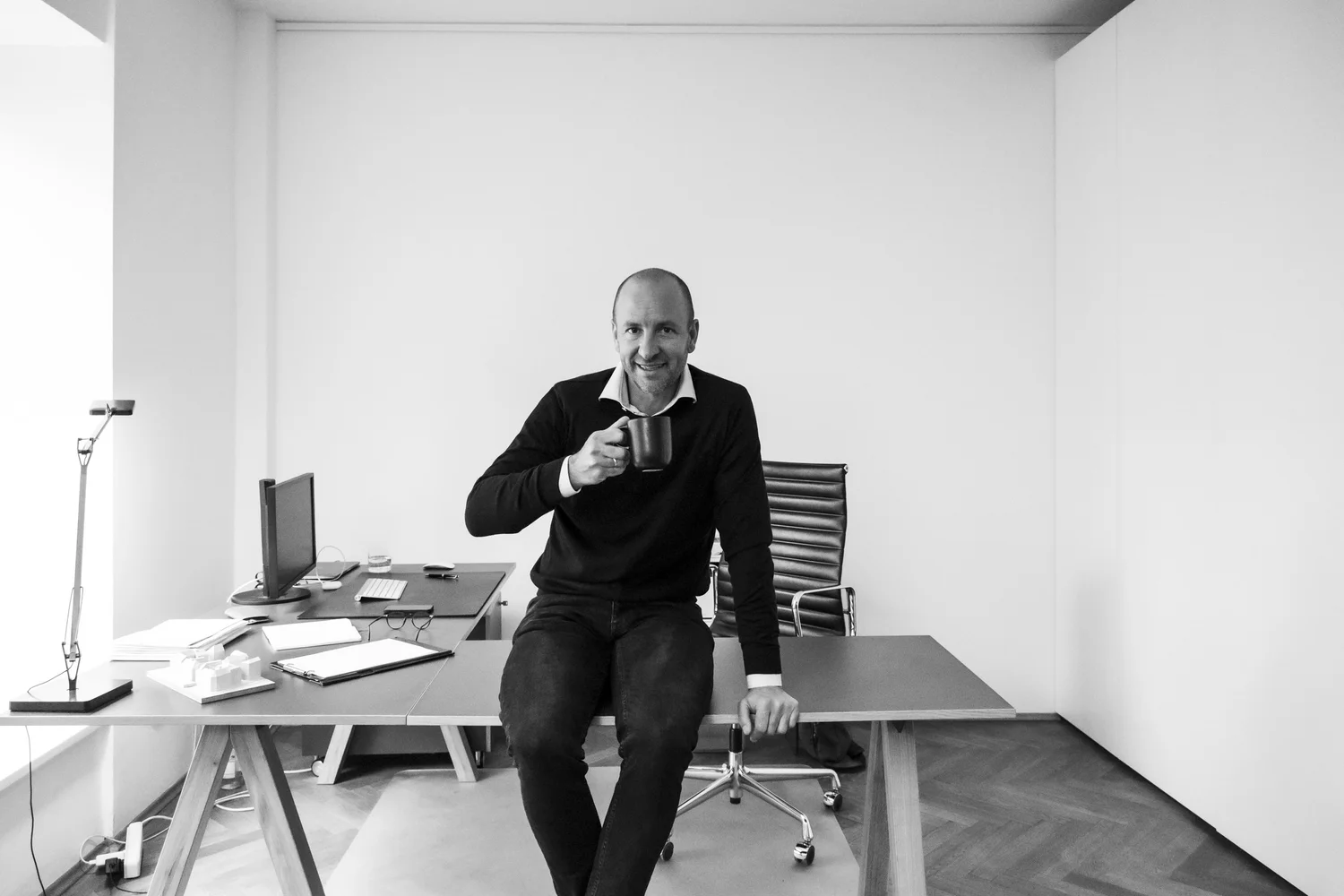 Architect of Büro Helmling sitting on his desk in a bright, minimalist white office, black-and-white business portrait — portrait by Janina Laszlo, portrait photographer in Munich