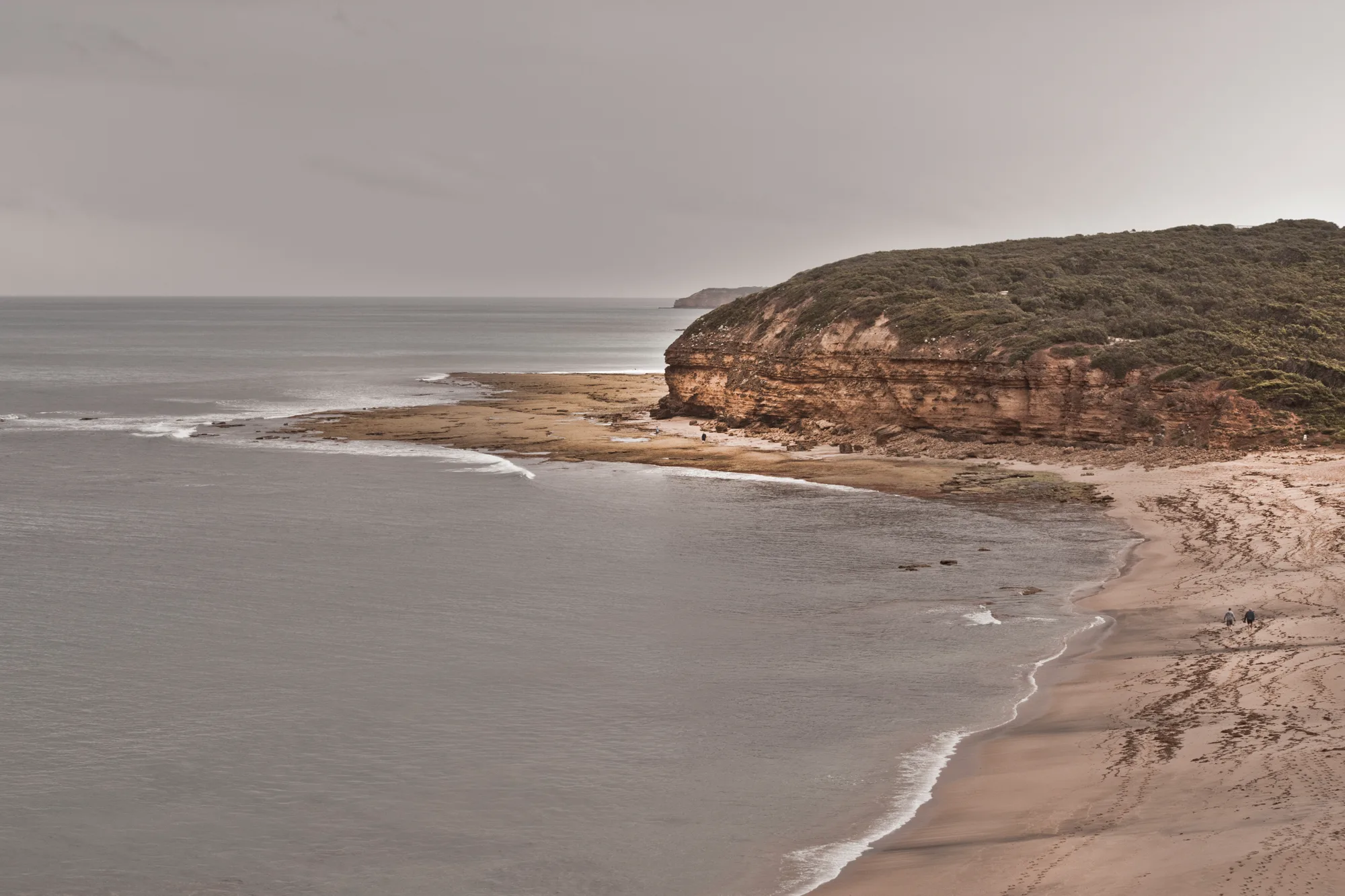 Sandstone cliff headland and a wide beach with two walkers under a grey sky — Australia, travel and fine art photography by Janina Laszlo