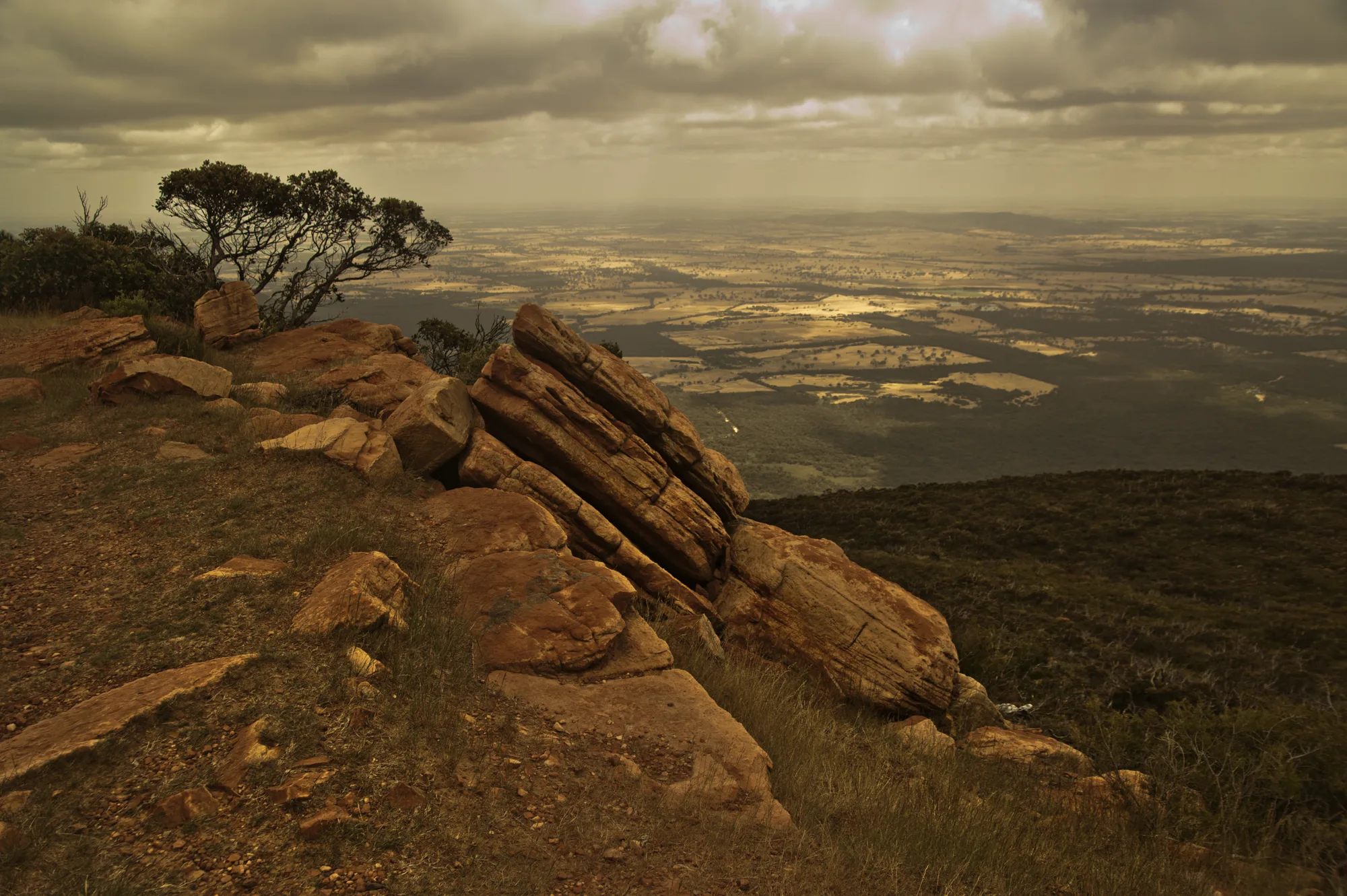 Tilted sandstone slabs on a summit in the Grampians above a patchwork of plains under dramatic clouds, Victoria — Australia, travel and fine art photography by Janina Laszlo