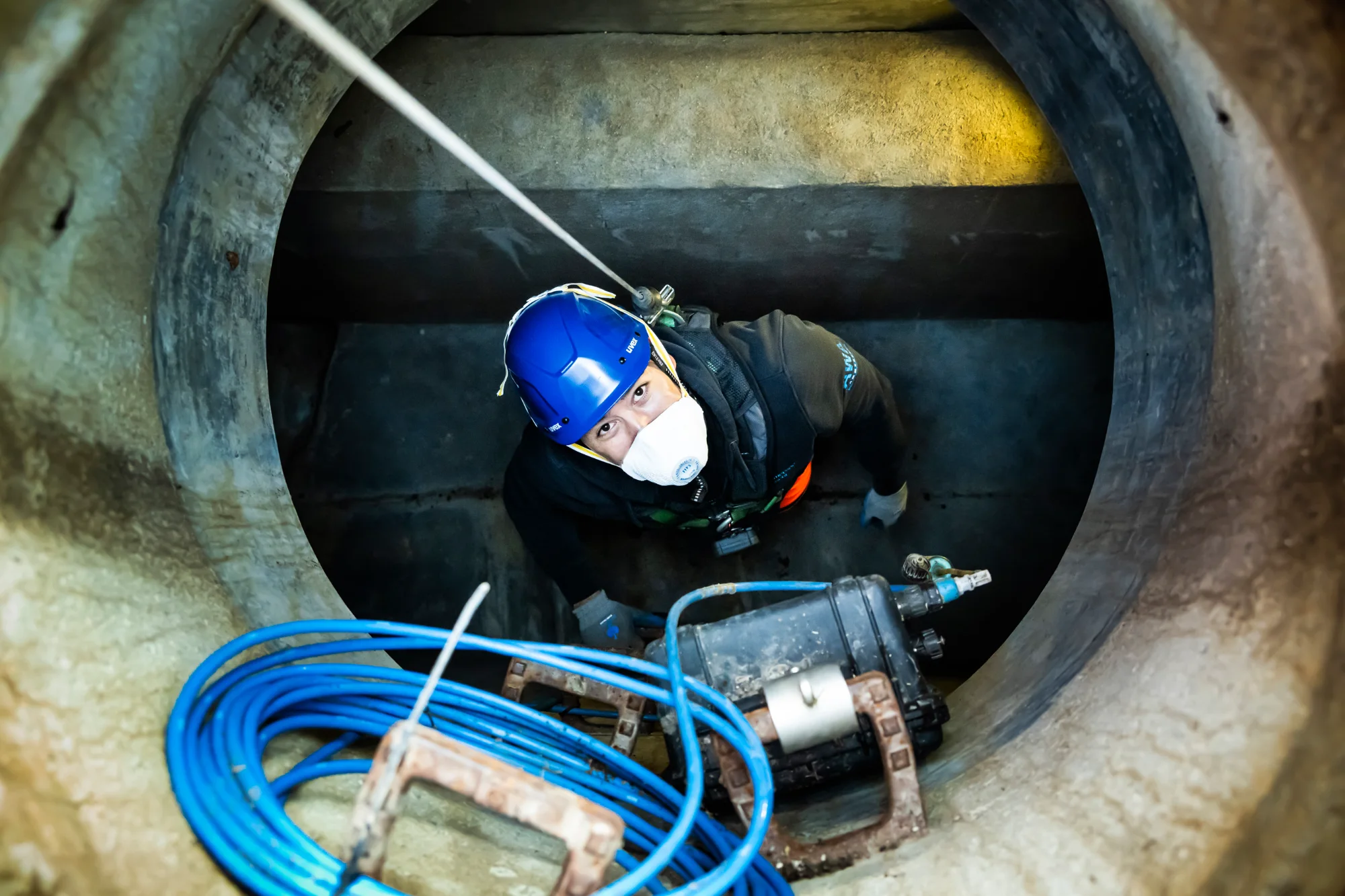 Worker in helmet and mask descending a manhole, seen from above, blue hoses beside — AWA Ammersee, industrial, corporate and portrait photography by Janina Laszlo