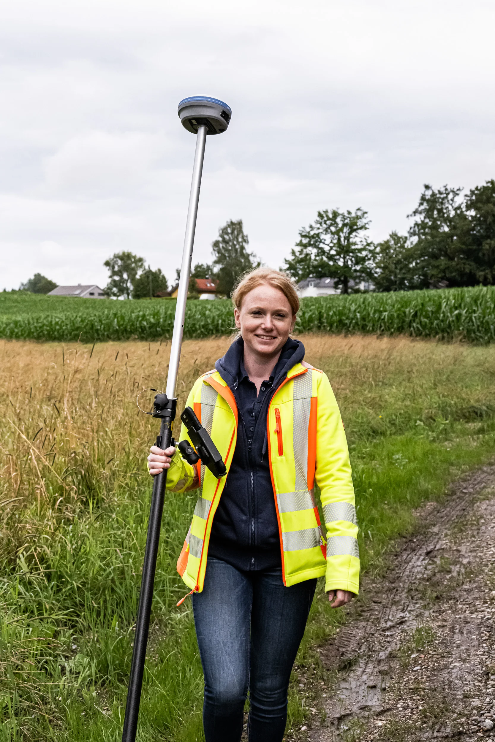 Surveyor in a hi-vis jacket with a GNSS rover pole at the edge of a maize field — AWA Ammersee, industrial, corporate and portrait photography by Janina Laszlo