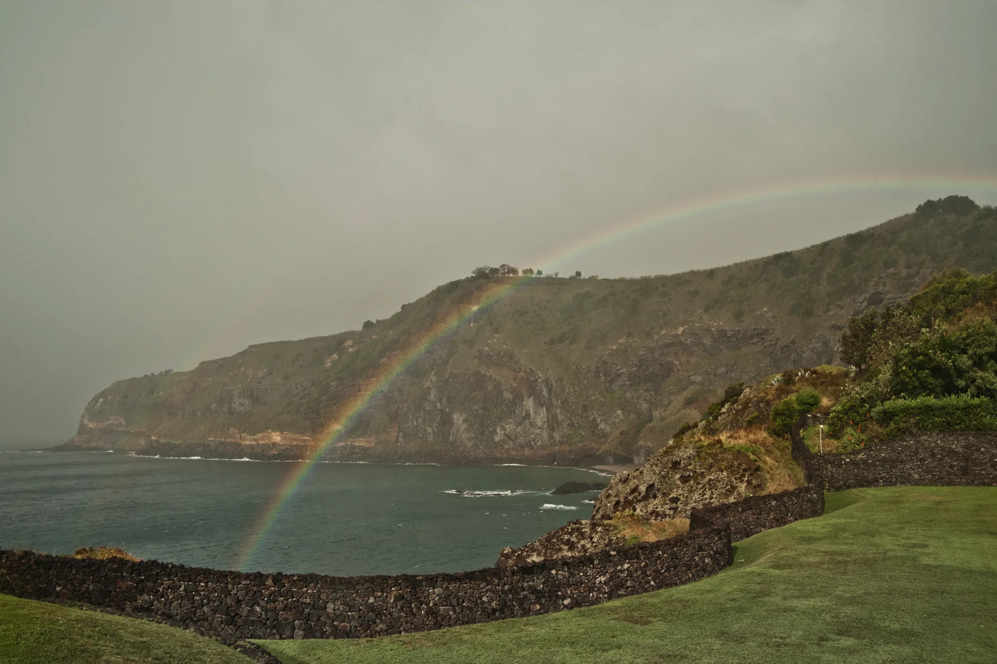Rainbow over green volcanic cliffs and a bay, lava-stone walls in the foreground — Azores, São Miguel, travel and fine art photography by Janina Laszlo