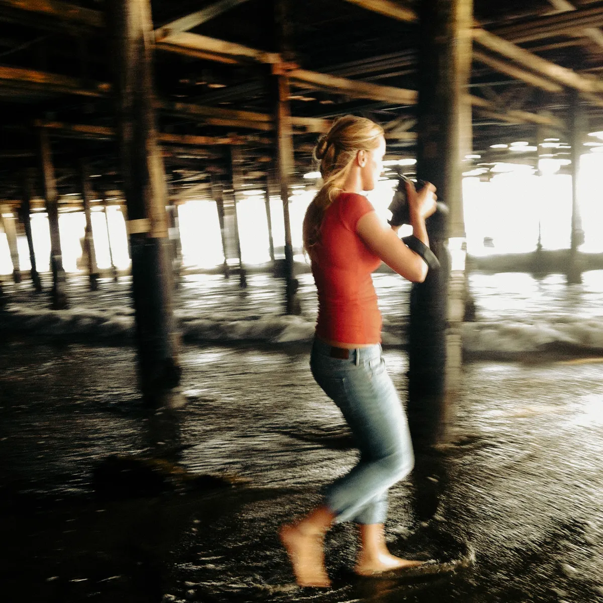 Photographer Janina Laszlo on set, wading barefoot under the Santa Monica Pier with her camera — behind the scenes with the German commercial and editorial photographer