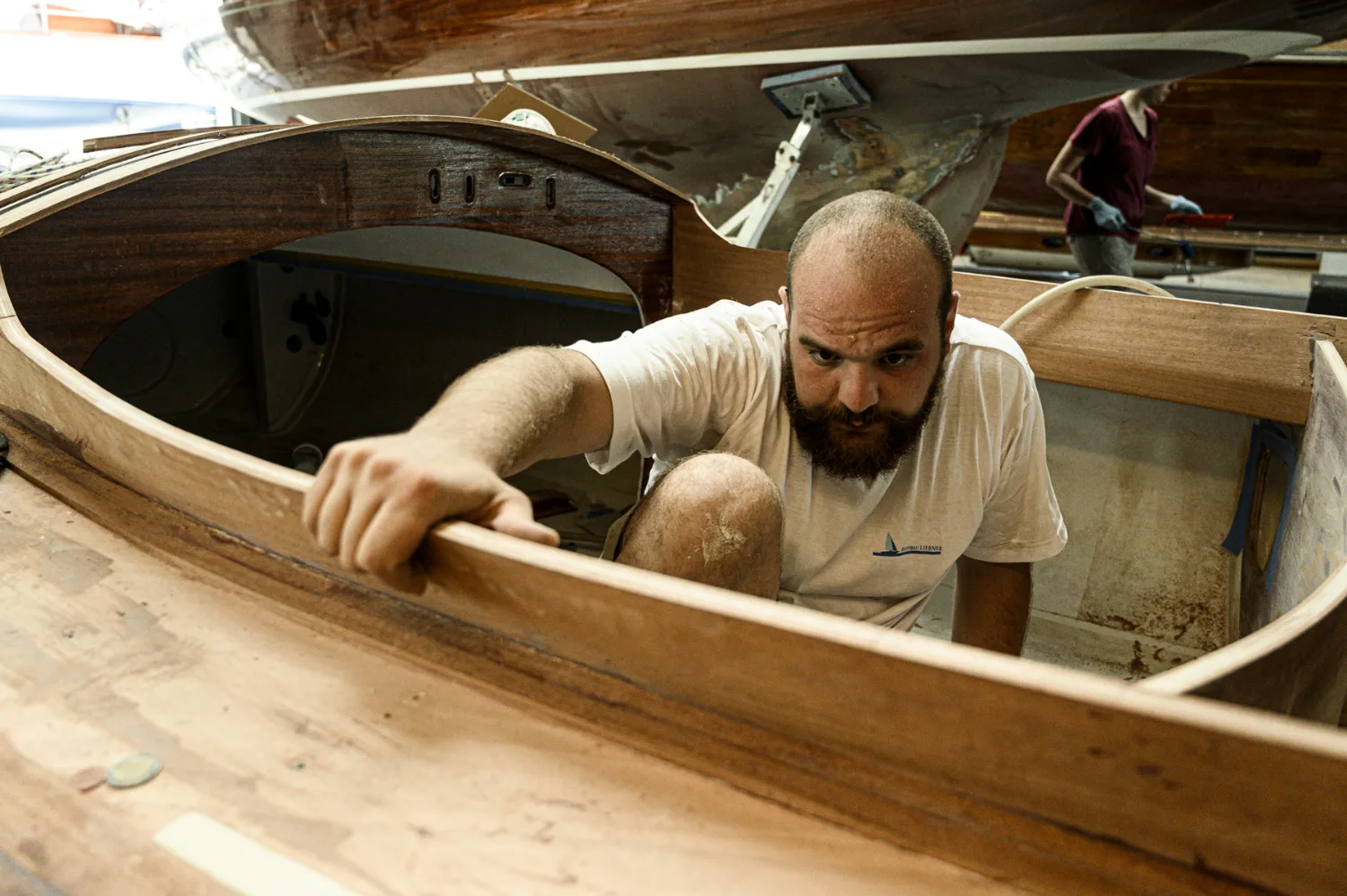 Boatbuilder crouching inside a mahogany cockpit, hand on the coaming, at Bootsbau Liebner — commercial and editorial photography by Janina Laszlo, photographer in Munich