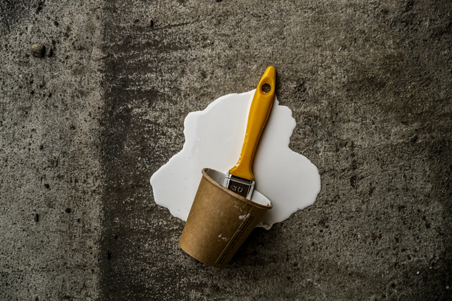 Paintbrush in a tipped paper cup with spilled white paint on concrete floor — commercial and editorial photography by Janina Laszlo, photographer in Munich