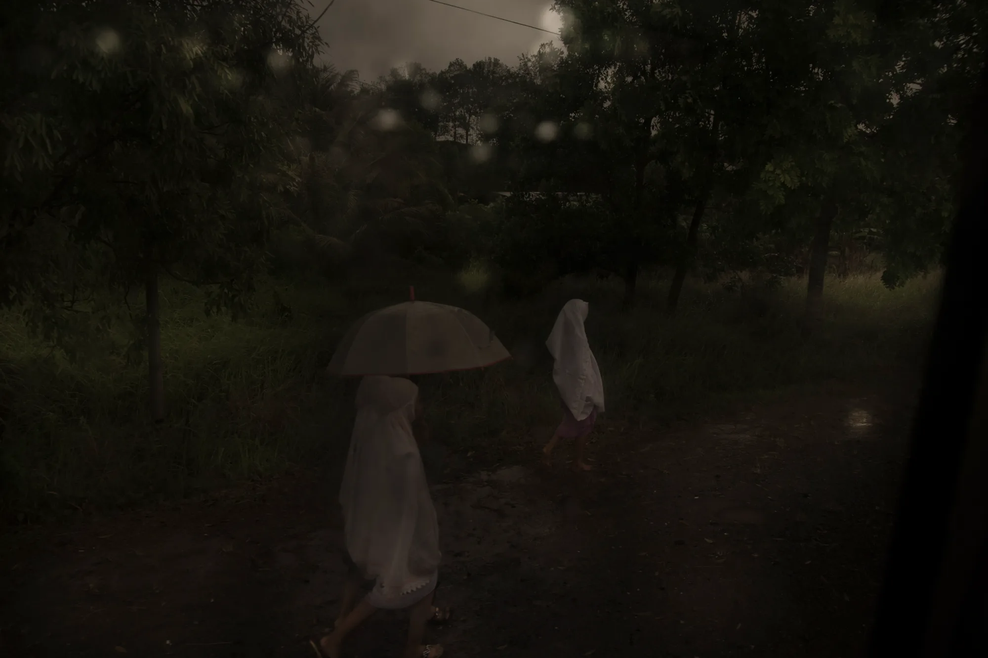Two children in white hooded capes walking a dark country road in the rain — Borneo & Pulau Mabul, documentary and travel photography by Janina Laszlo