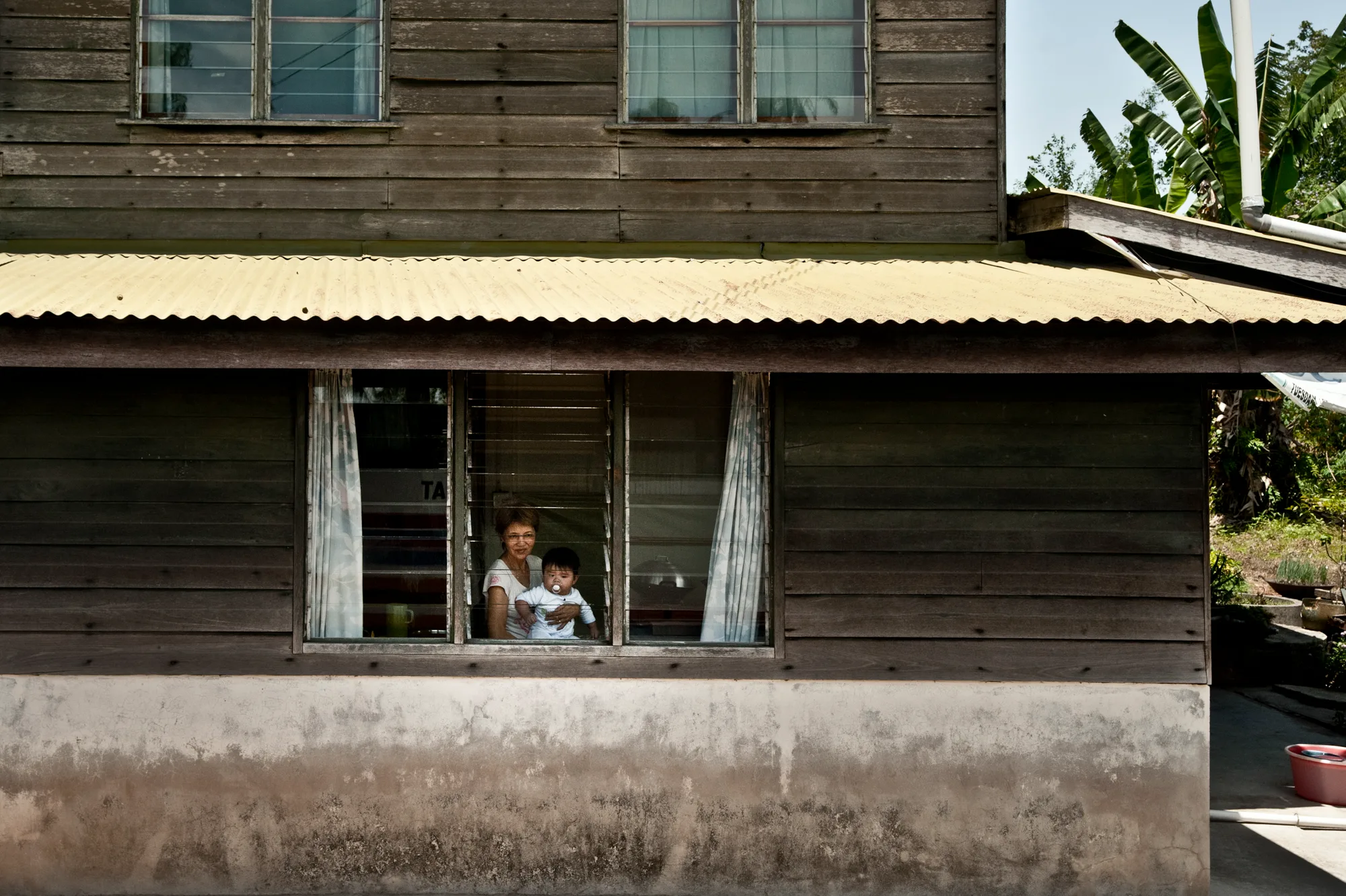 Grandmother holding a baby at the window of a wooden house — Borneo & Pulau Mabul, documentary and travel photography by Janina Laszlo