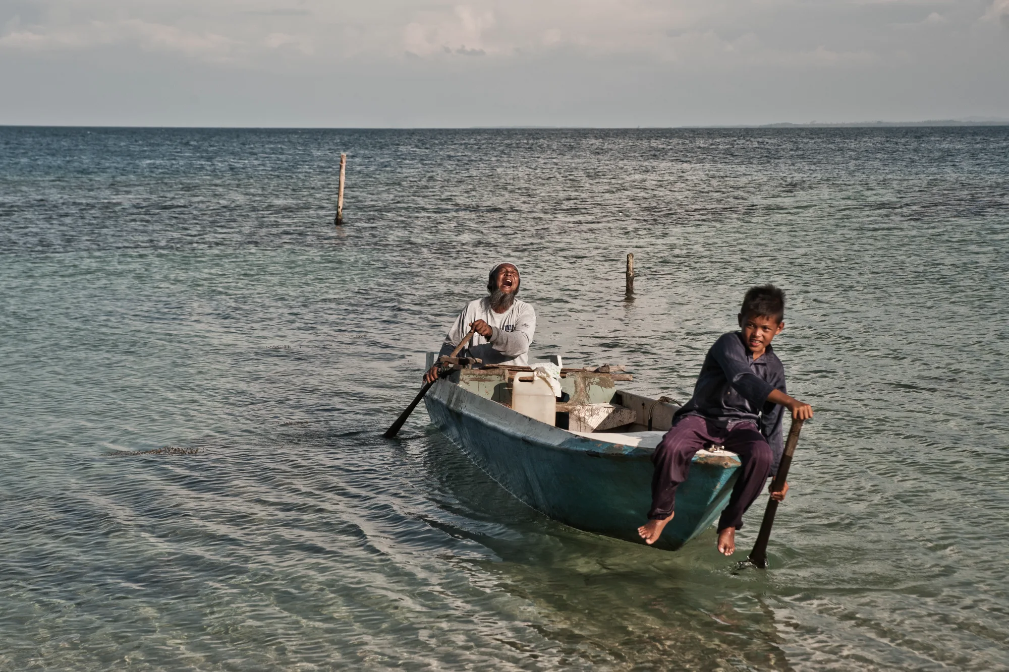 Laughing fisherman rowing a small boat while a boy sits on the bow — Borneo & Pulau Mabul, documentary and travel photography by Janina Laszlo