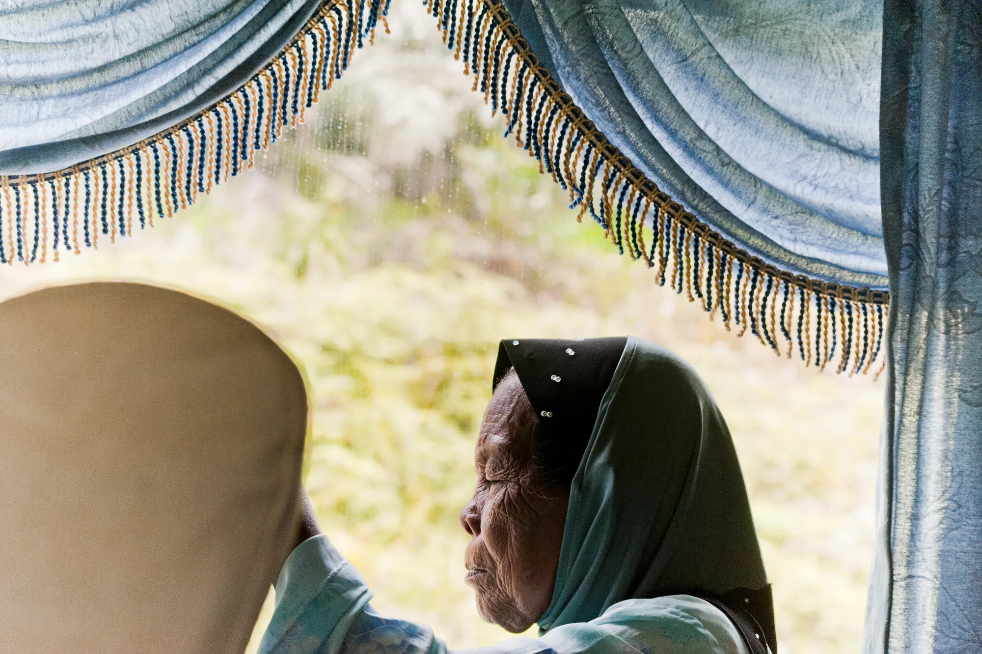 Elderly woman in a headscarf by a curtained bus window — Borneo & Pulau Mabul, documentary and travel photography by Janina Laszlo