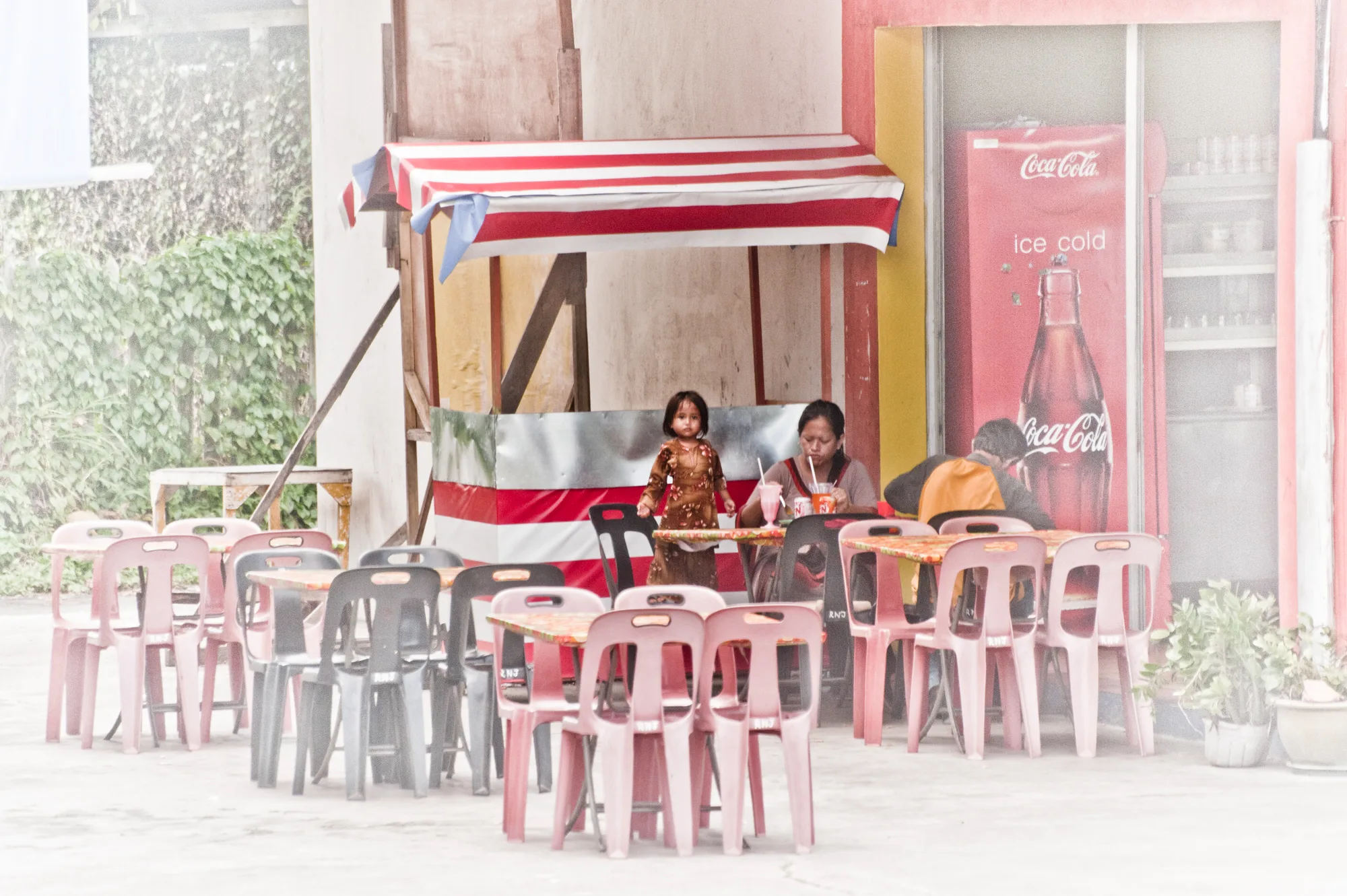 Girl standing at a food stall with pink plastic chairs and a Coca-Cola fridge — Borneo & Pulau Mabul, documentary and travel photography by Janina Laszlo