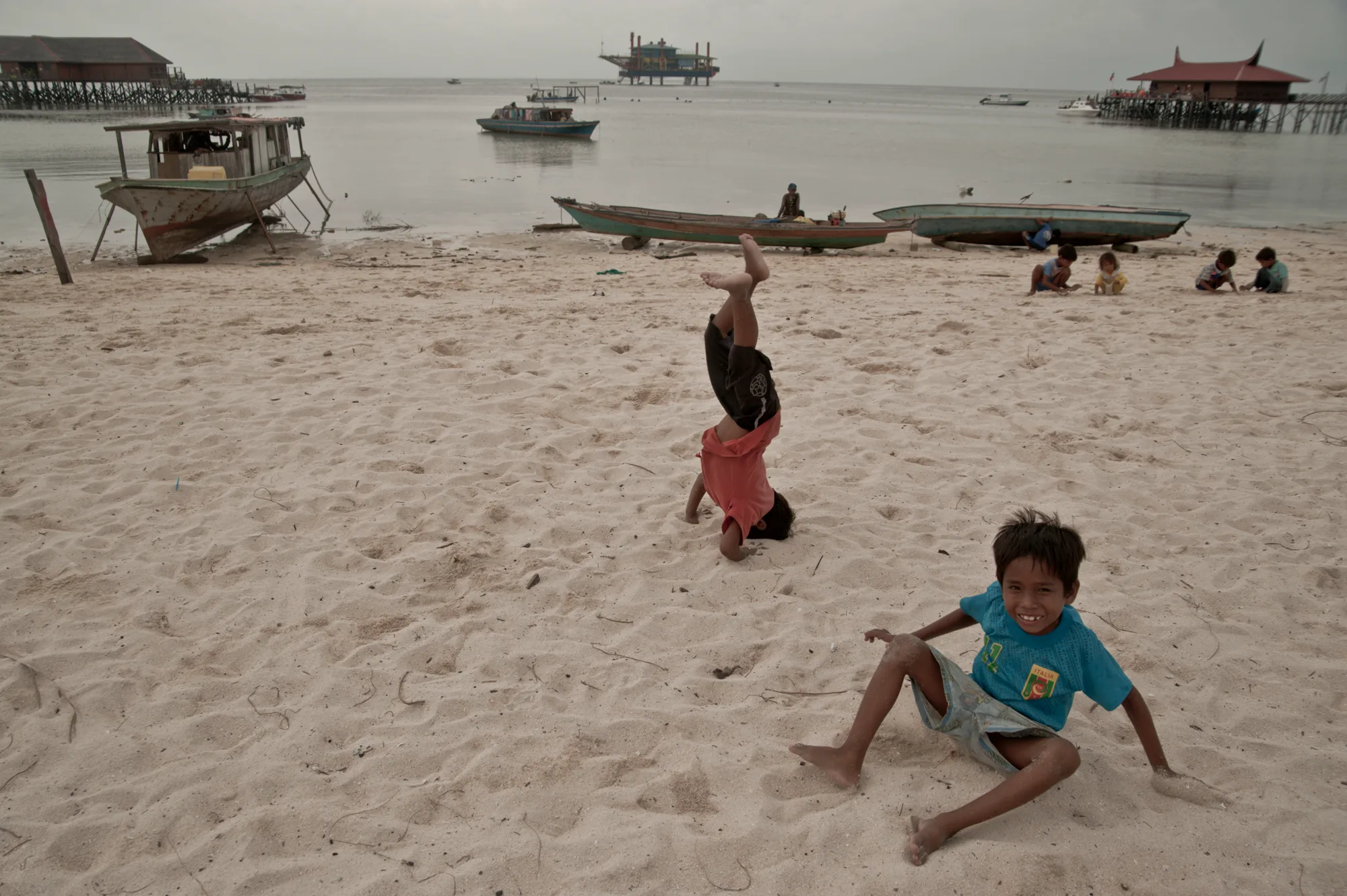 Boy doing a headstand on the beach with boats and a former oil rig dive resort beyond, Pulau Mabul, Sabah — Borneo & Pulau Mabul, documentary and travel photography by Janina Laszlo