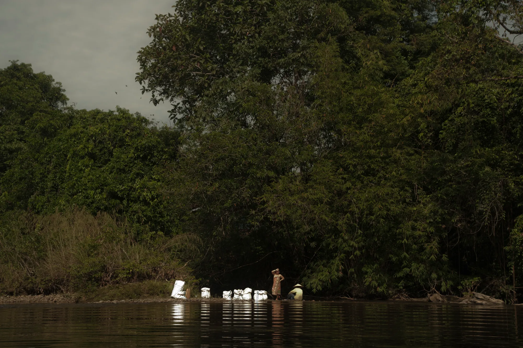 Two women with white sacks on a riverbank below tall rainforest trees — Borneo & Pulau Mabul, documentary and travel photography by Janina Laszlo