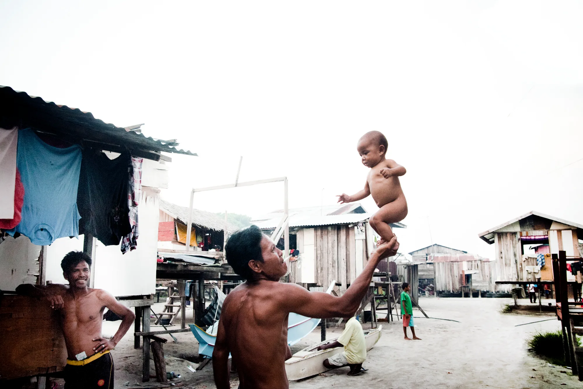 Man balancing a baby on his hand in a village while others watch — Borneo & Pulau Mabul, documentary and travel photography by Janina Laszlo