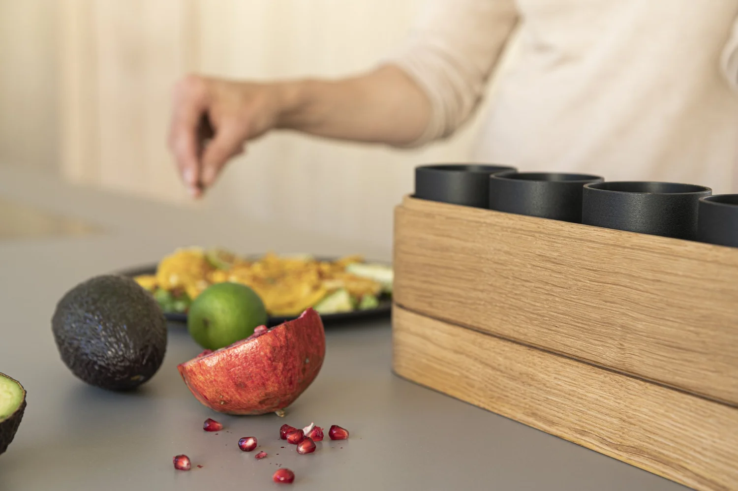 Hand placing pomegranate and avocado next to an oak bulthaup kitchen box with black storage tubs — campaign photography by Janina Laszlo, Munich