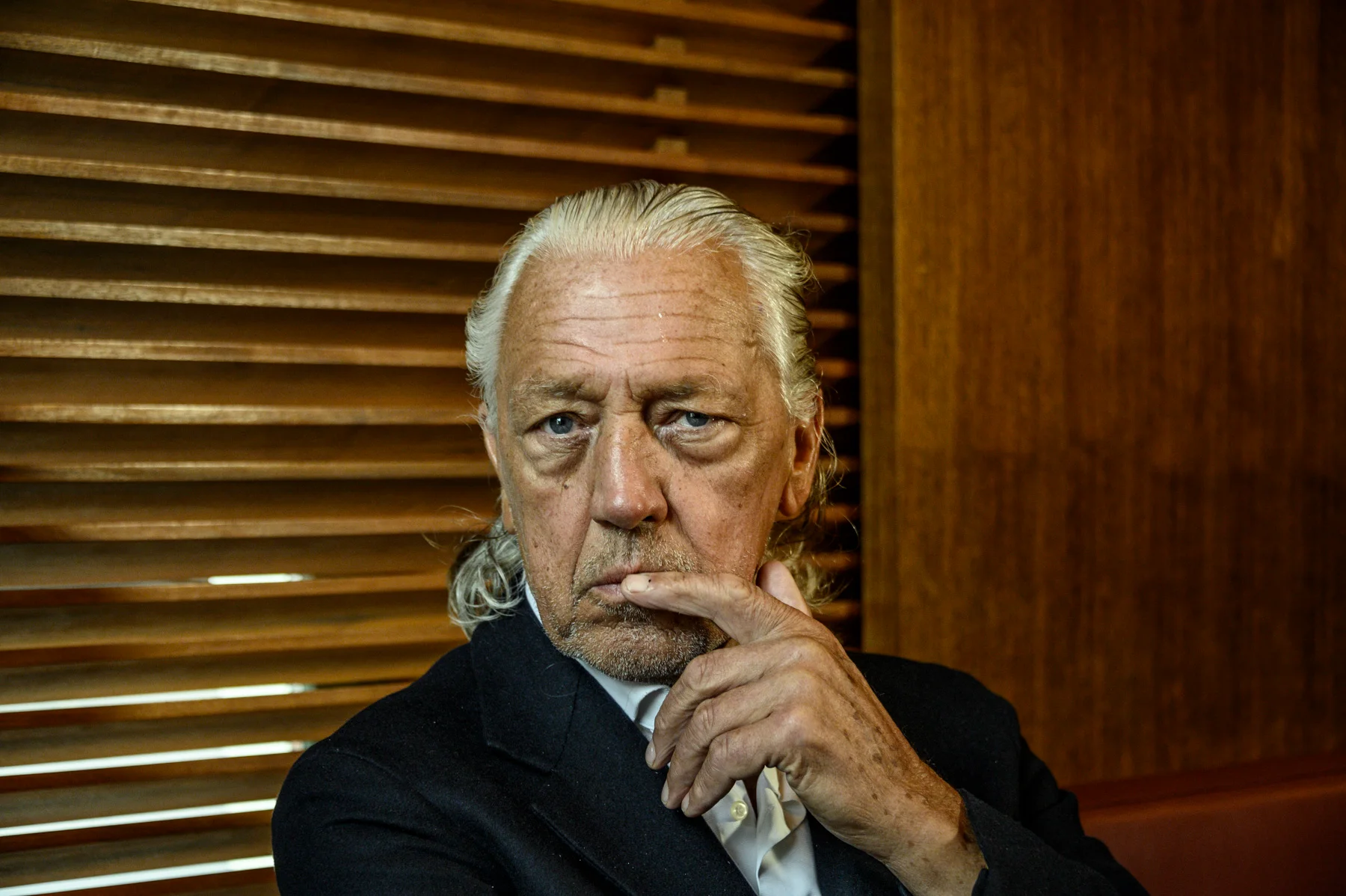 Charles Schumann with his hand at his chin in front of wooden blinds — Charles Schumann at Schumann’s Bar, Munich, editorial portrait photography by Janina Laszlo