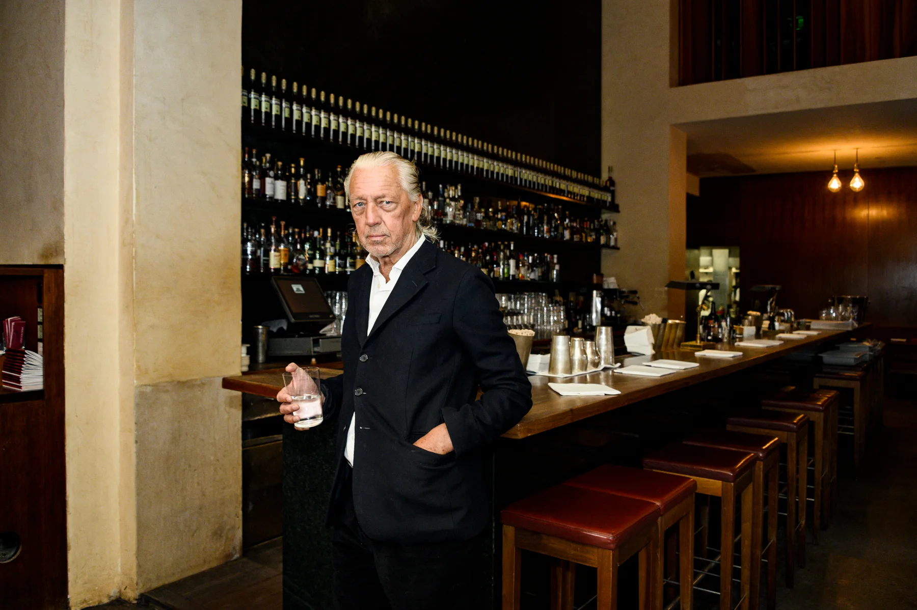 Charles Schumann with a glass of water at the bar of Schumann’s — Charles Schumann at Schumann’s Bar, Munich, editorial portrait photography by Janina Laszlo