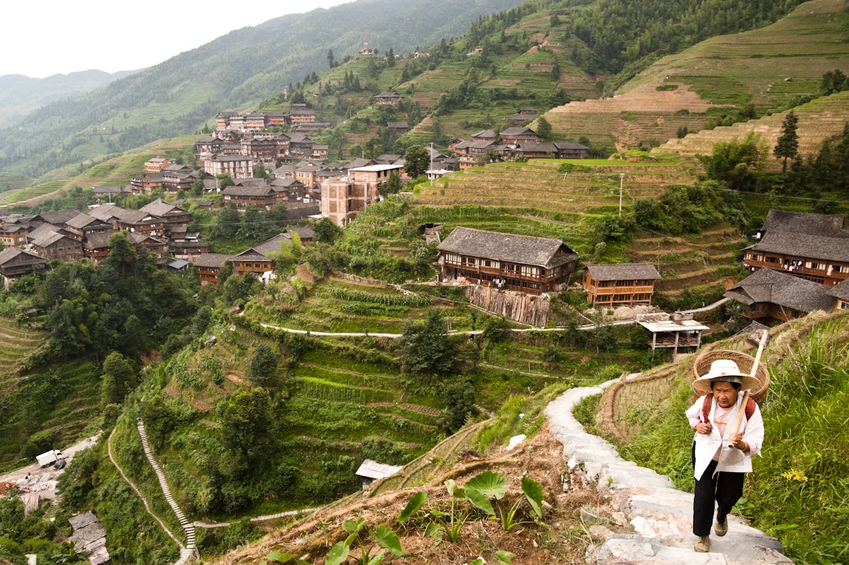 Elderly woman with a basket climbing a stone path above a village of wooden houses and rice terraces, Guangxi — China, documentary and travel photography by Janina Laszlo