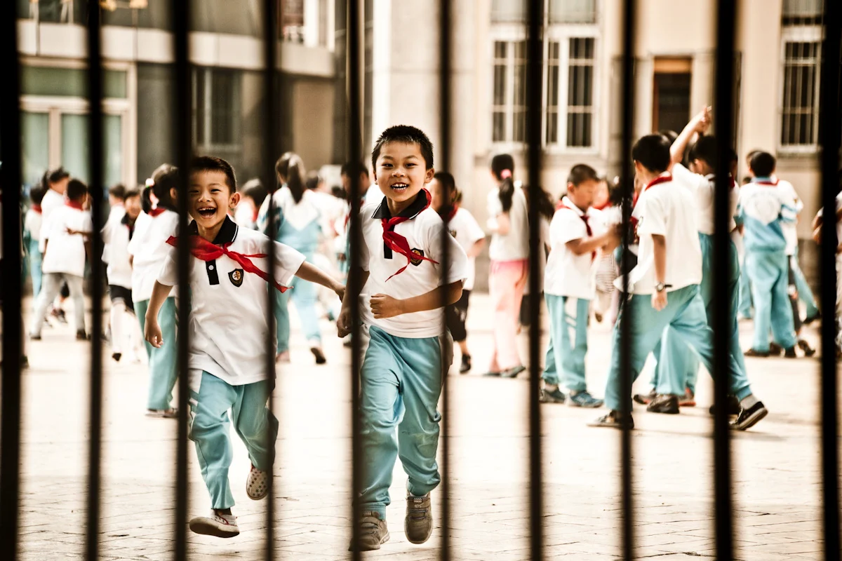 Laughing schoolchildren with red scarves running behind a fence, Qingdao — China, documentary and travel photography by Janina Laszlo