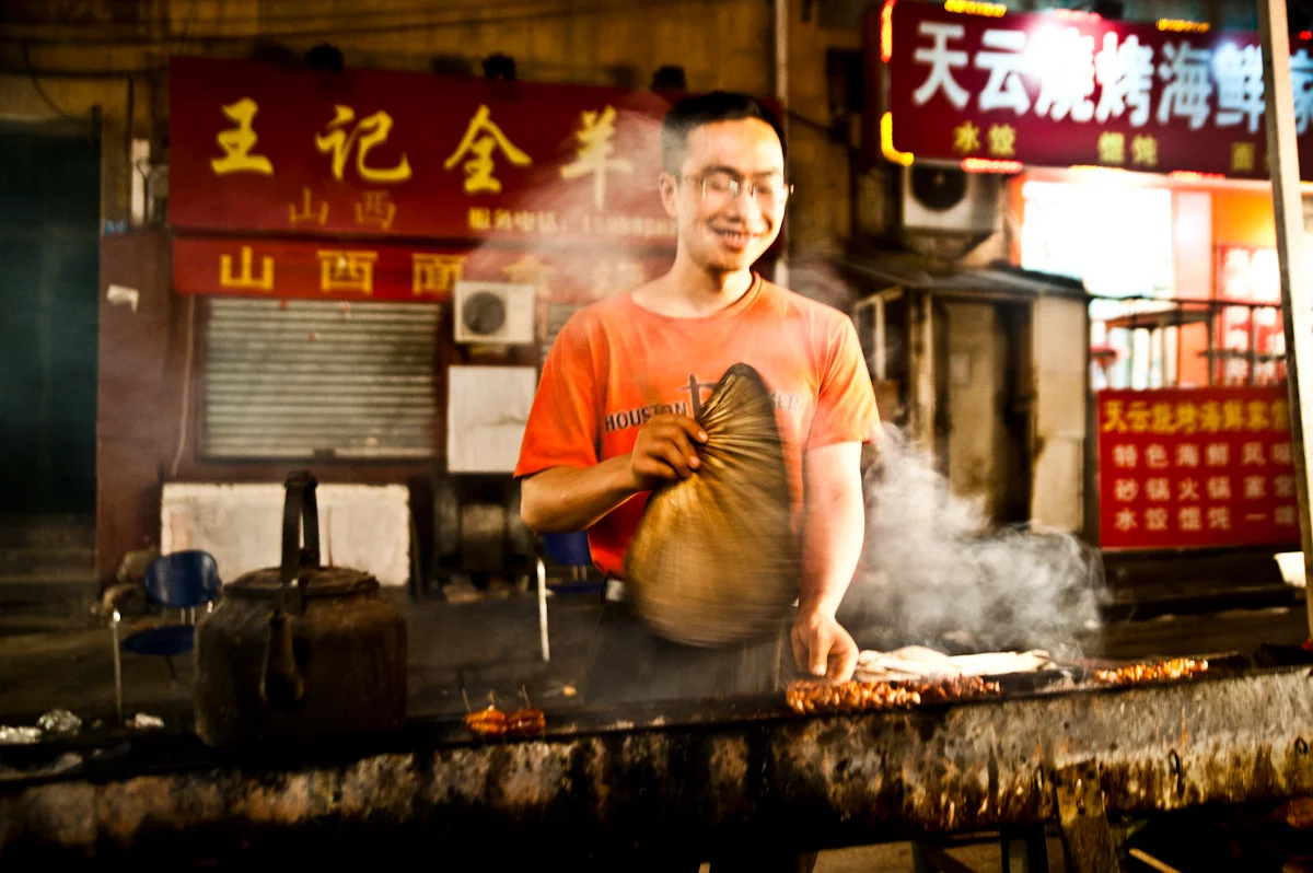 Grill cook fanning lamb skewers at a night street stall, Qingdao — China, documentary and travel photography by Janina Laszlo