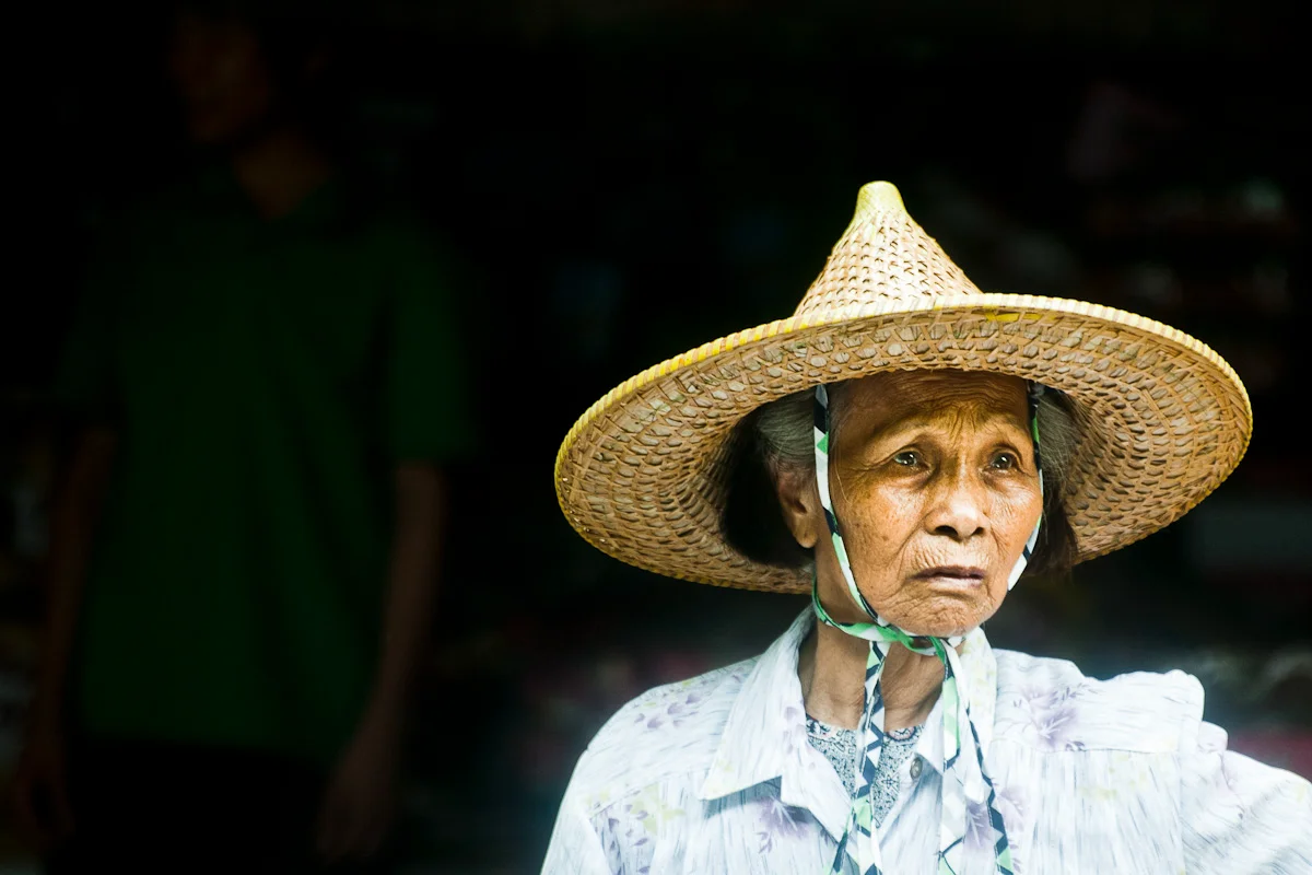 Elderly woman in a conical straw hat against a dark doorway — China, documentary and travel photography by Janina Laszlo