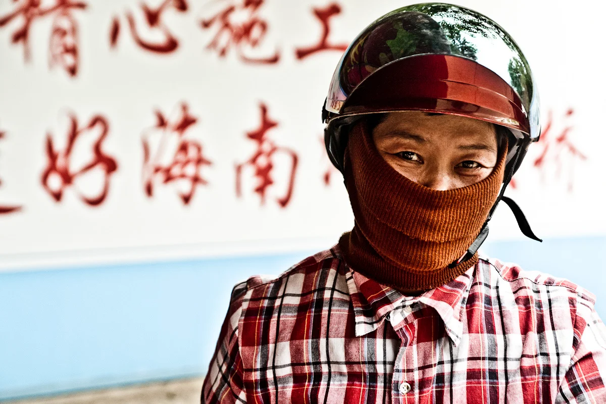 Woman in a motorbike helmet and knitted face mask before a red slogan mentioning Hainan — China, documentary and travel photography by Janina Laszlo