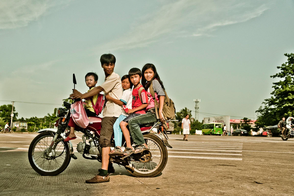 Father with four children on one motorbike at a wide crossroads, Hainan — China, documentary and travel photography by Janina Laszlo
