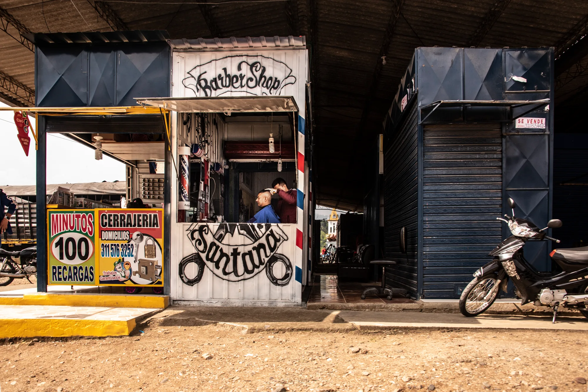 Barber cutting hair inside a small painted barbershop kiosk next to a parked motorbike — Colombia, travel and documentary photography by Janina Laszlo