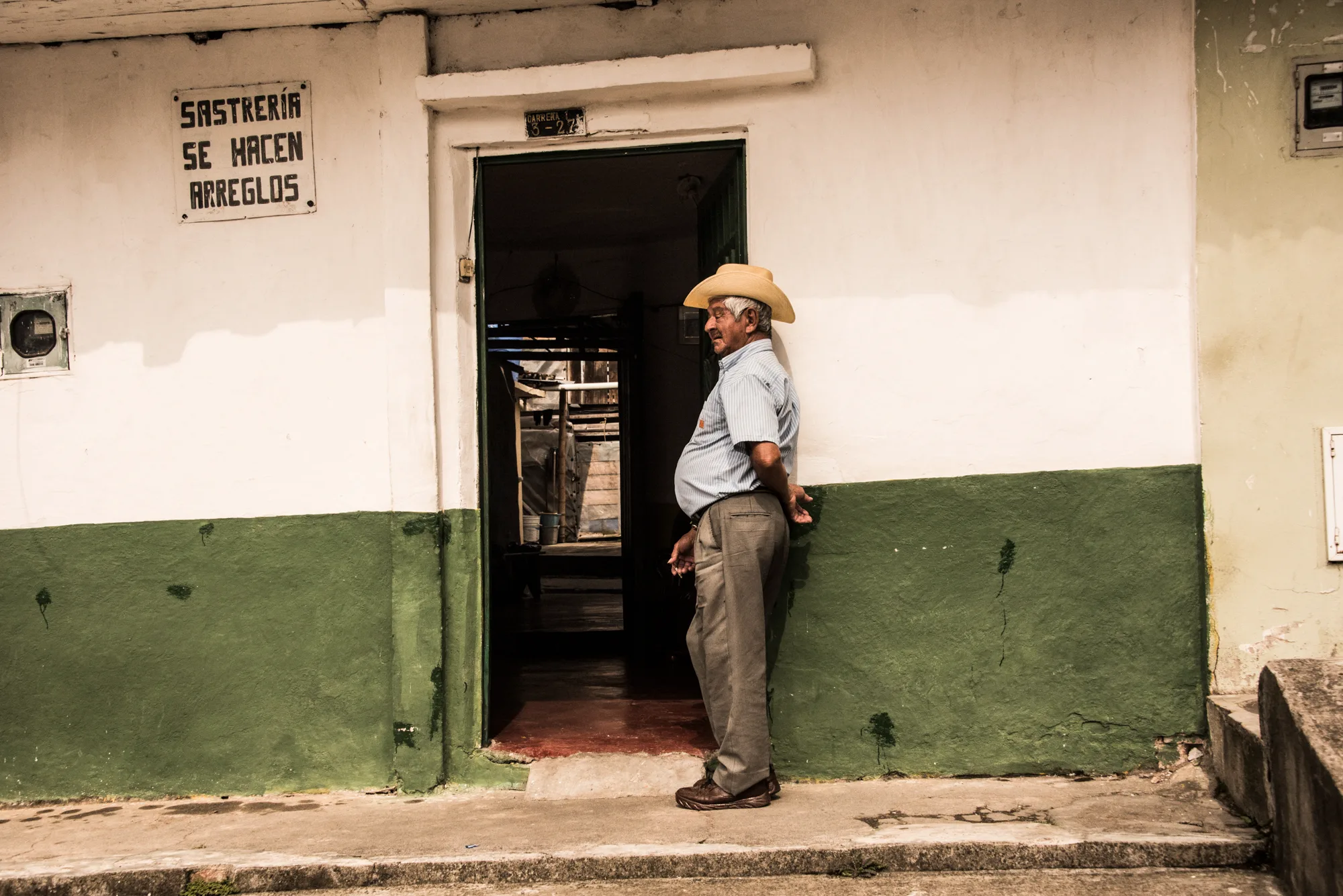Elderly man in a straw hat leaning by the doorway of a tailor shop with a green-and-white facade — Colombia, travel and documentary photography by Janina Laszlo