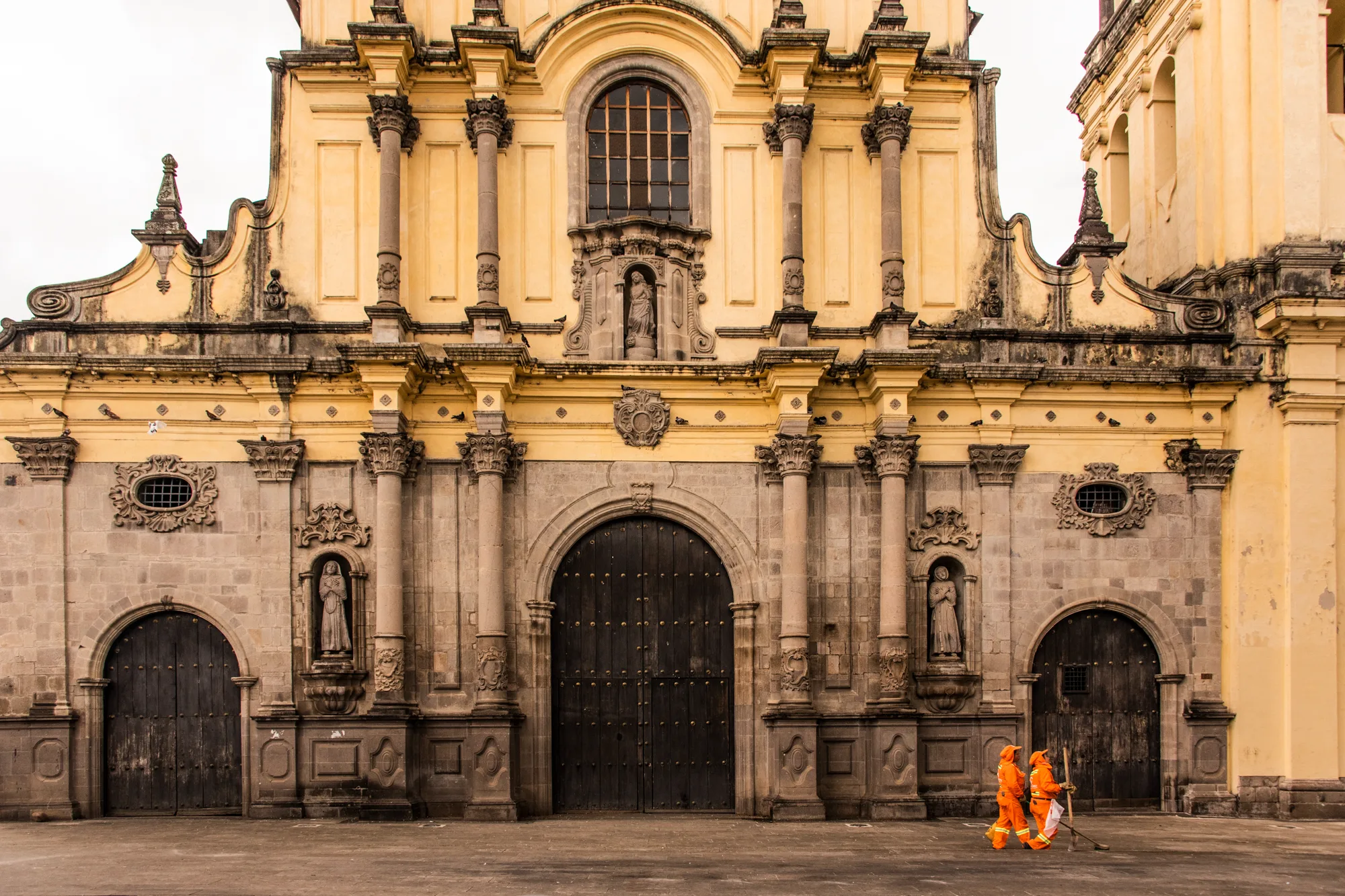 Two street sweepers in orange walking past the facade of a yellow colonial church — Colombia, travel and documentary photography by Janina Laszlo