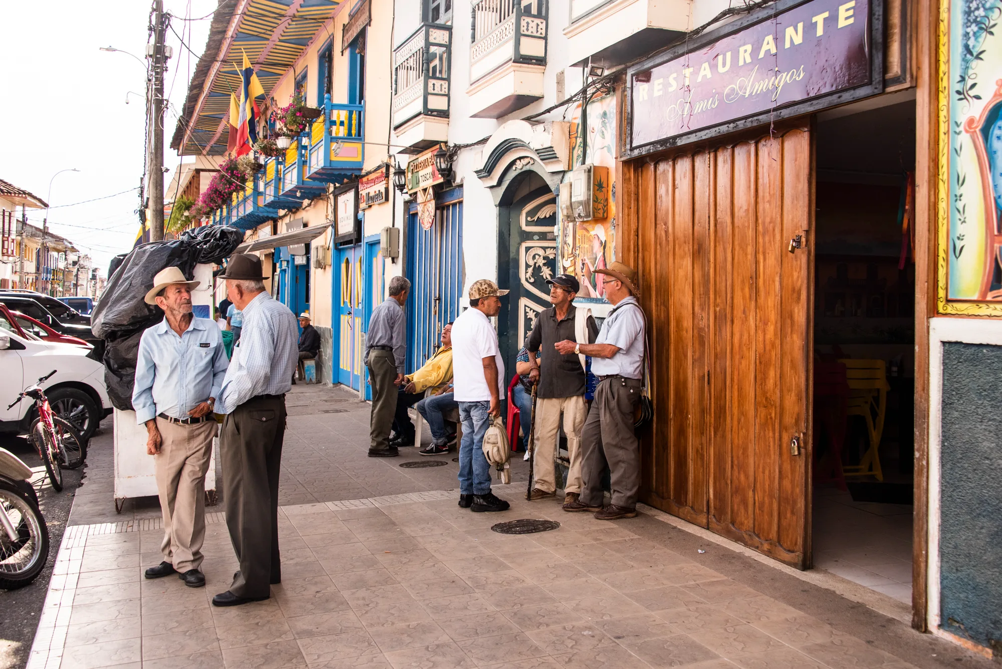 Elderly men in hats chatting outside a restaurant on a colourful street, Jardín, Antioquia — Colombia, travel and documentary photography by Janina Laszlo