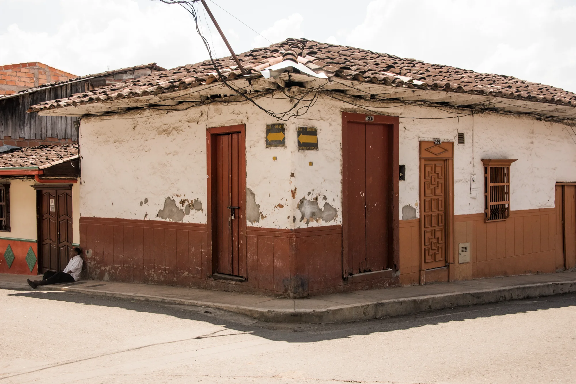 Weathered white corner house with red doors and a man resting on the pavement — Colombia, travel and documentary photography by Janina Laszlo