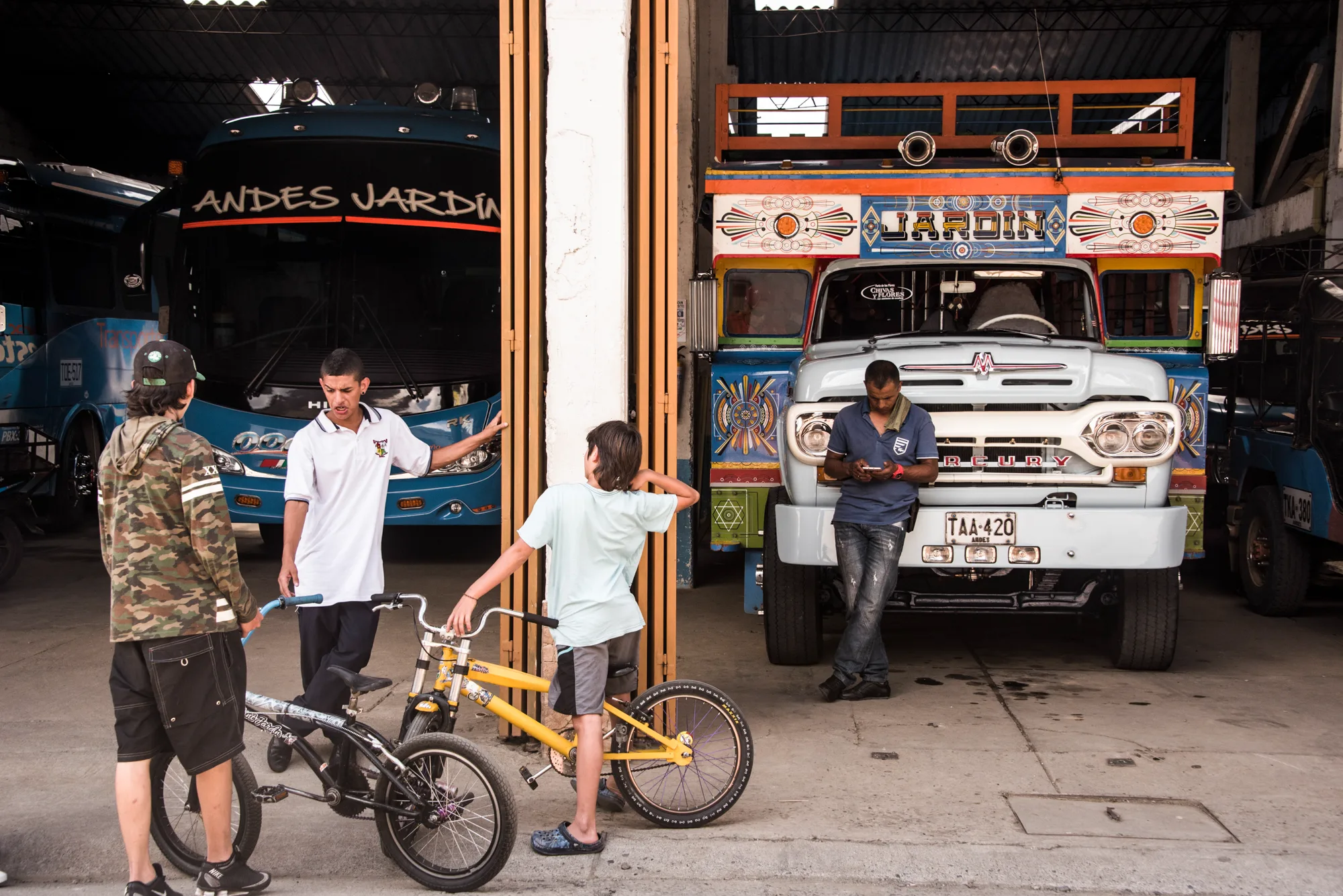 Boys with bikes in front of a blue bus and a painted chiva truck in a bus depot, Jardín, Antioquia — Colombia, travel and documentary photography by Janina Laszlo