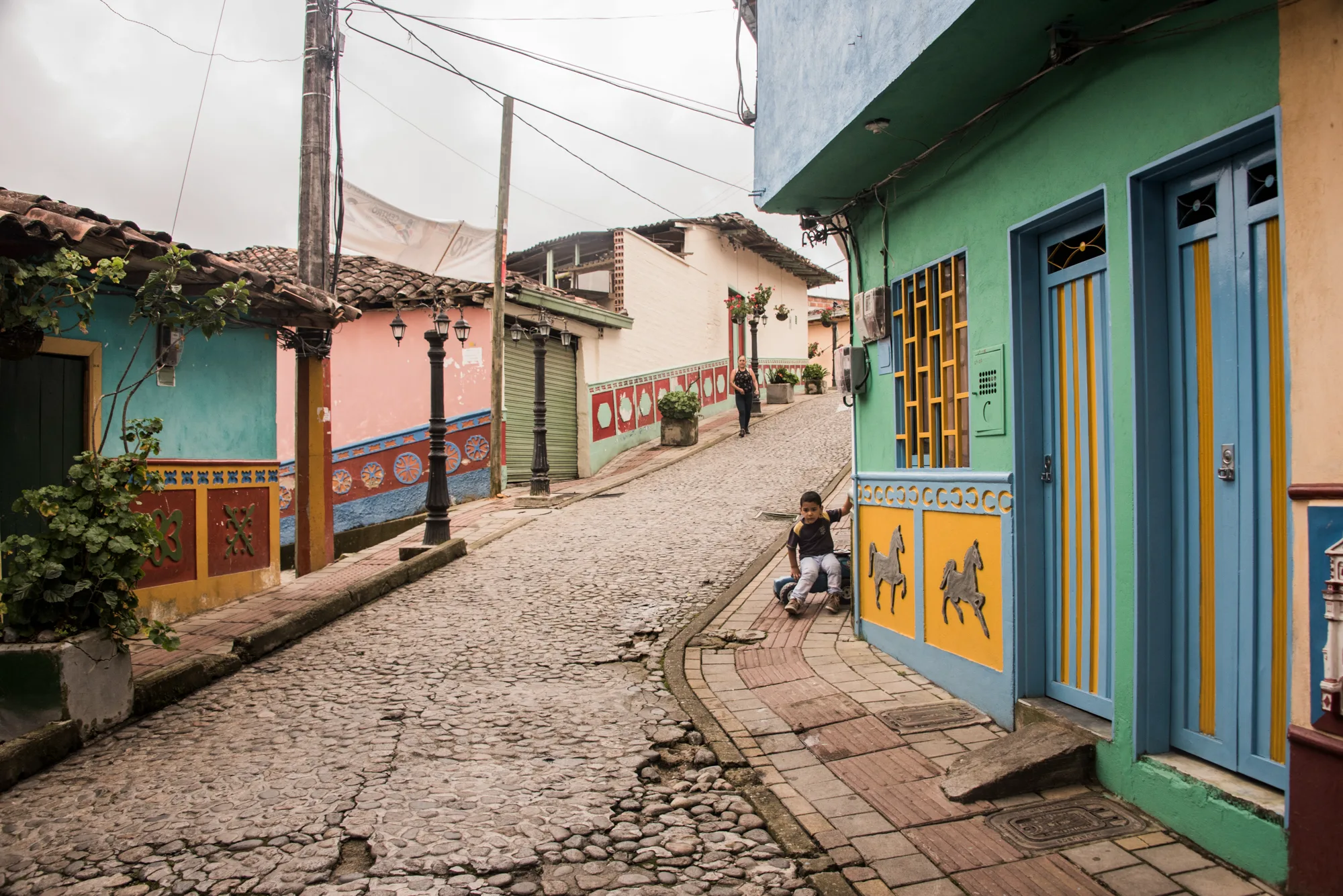 Cobbled lane with colourful houses and painted zócalos, a boy sliding down the pavement, Guatapé — Colombia, travel and documentary photography by Janina Laszlo