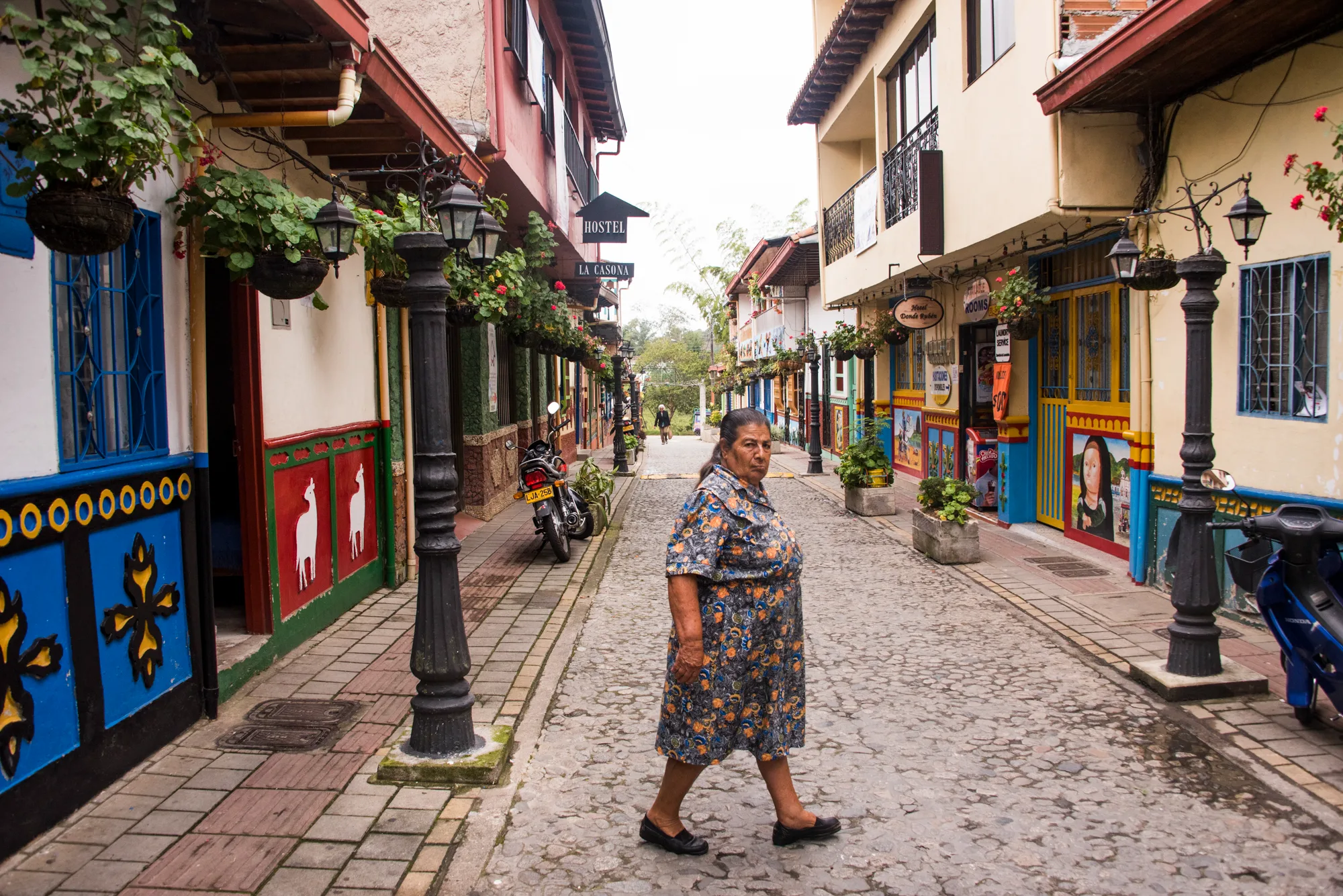 Elderly woman in a floral dress crossing a colourful cobbled street, Guatapé — Colombia, travel and documentary photography by Janina Laszlo