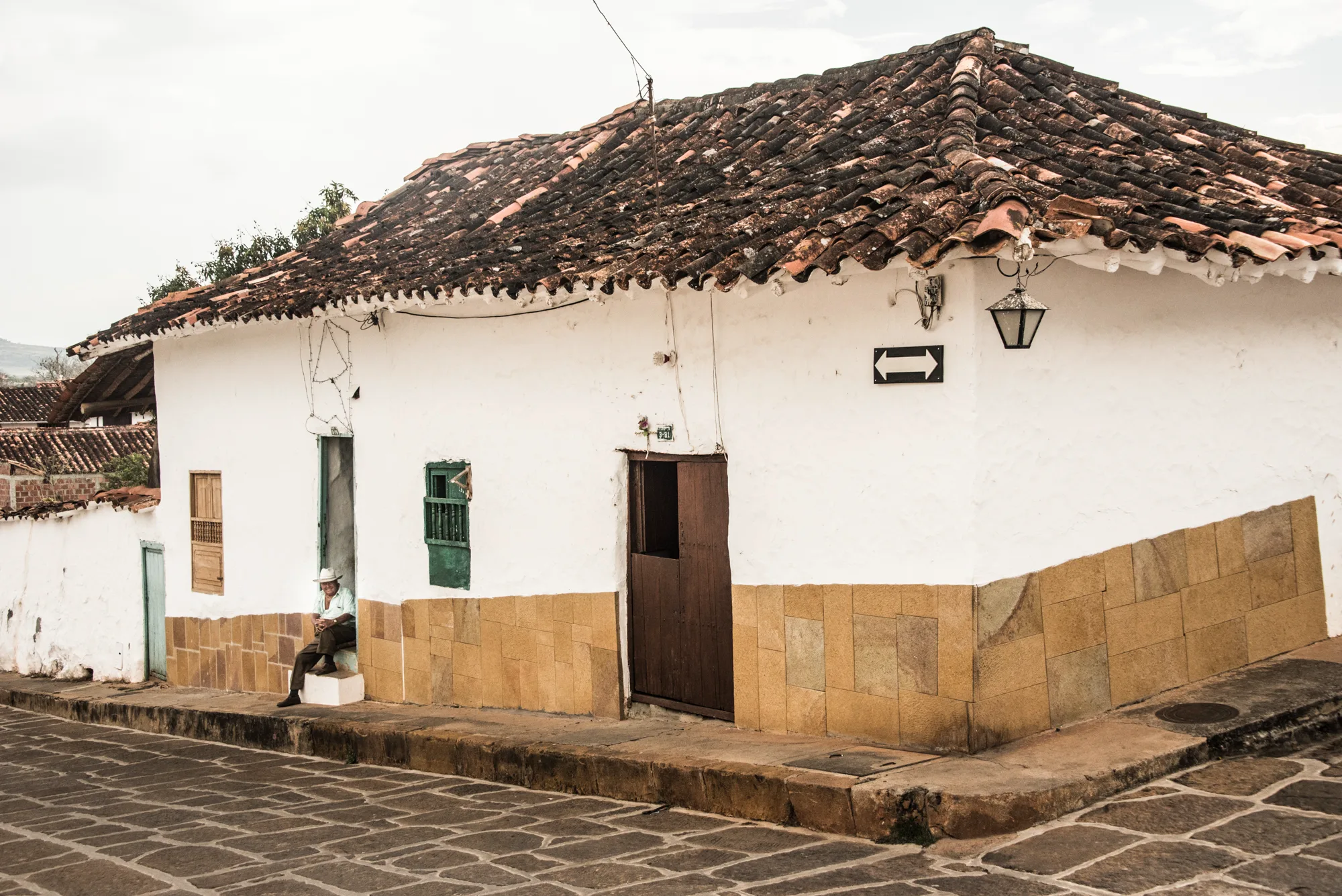 Man in a white hat sitting in the doorway of a white colonial corner house, Barichara — Colombia, travel and documentary photography by Janina Laszlo