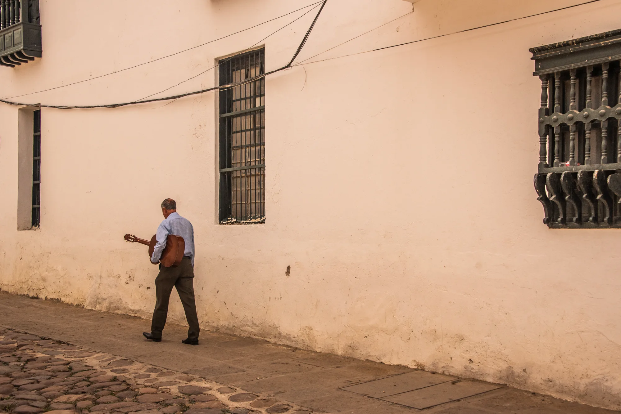 Man carrying a guitar walking along a pale pink colonial wall, Villa de Leyva — Colombia, travel and documentary photography by Janina Laszlo