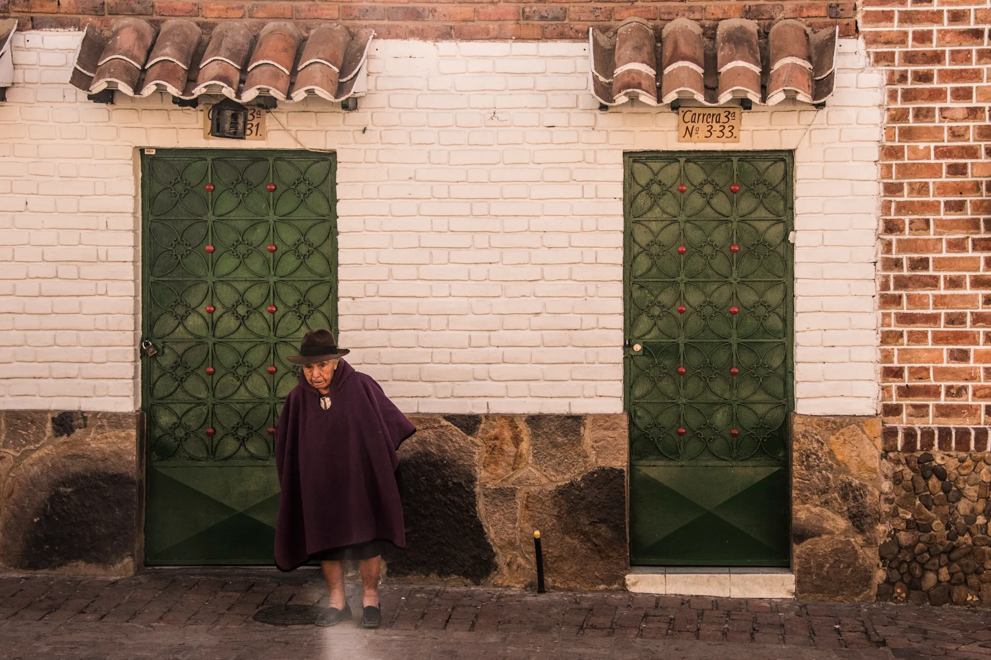 Elderly woman in a purple ruana and hat in front of green iron doors — Colombia, travel and documentary photography by Janina Laszlo