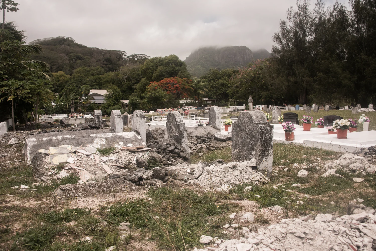 Weathered old graves and freshly whitewashed tombs with flowers below a cloud-capped peak, Rarotonga — Cook Islands, travel photography by Janina Laszlo
