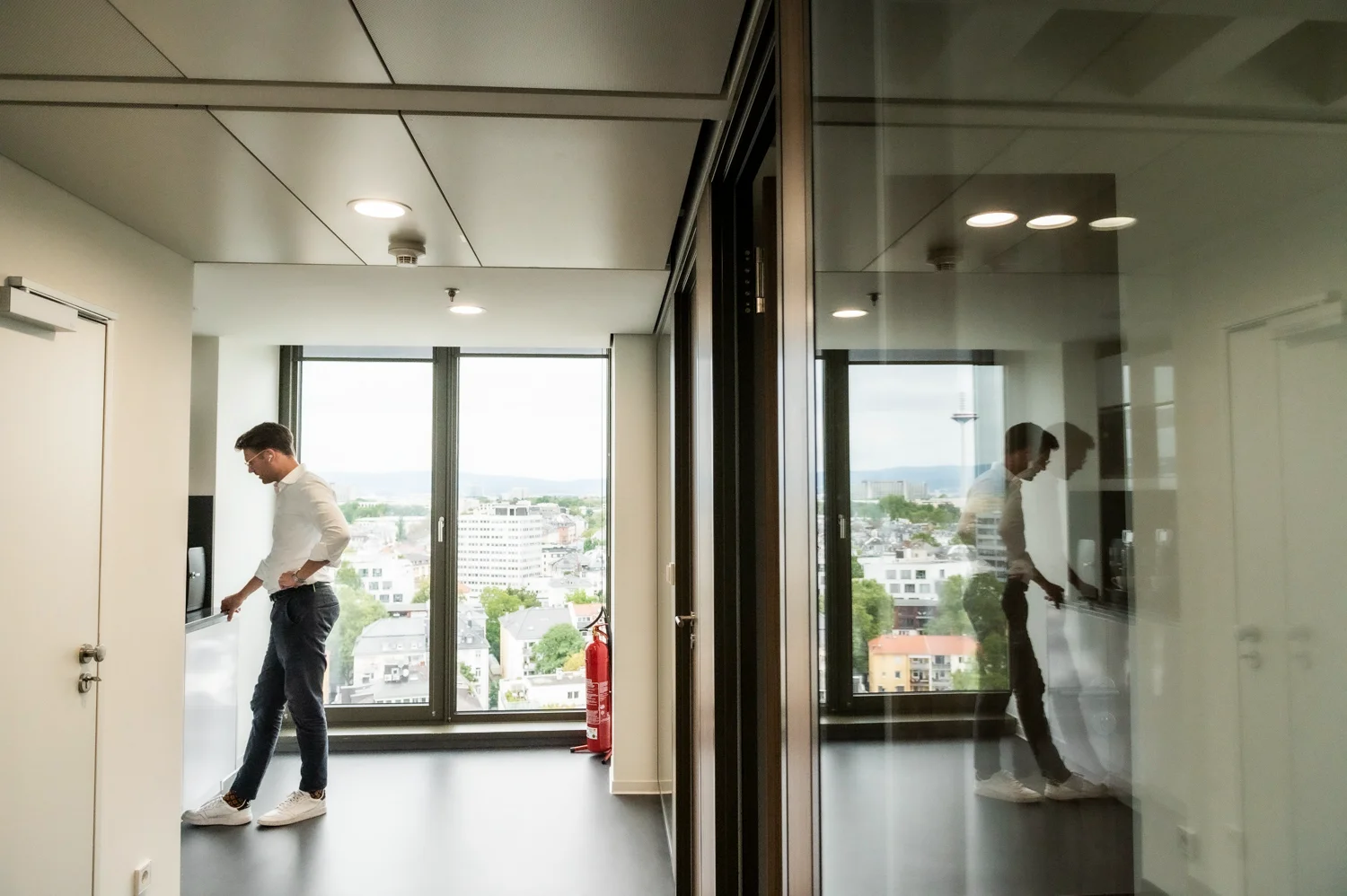 Employee in a white shirt at the office kitchenette, mirrored in a glass wall, with a view over Frankfurt am Main — Digital+, corporate photography by Janina Laszlo