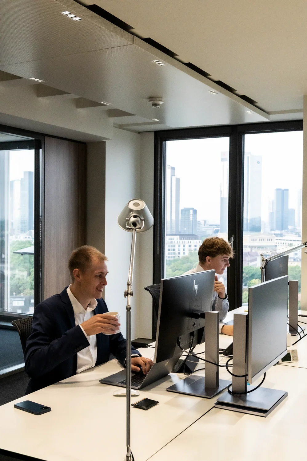 Two colleagues at their desks with monitors in front of floor-to-ceiling windows and the Frankfurt skyline, Frankfurt am Main — Digital+, corporate photography by Janina Laszlo