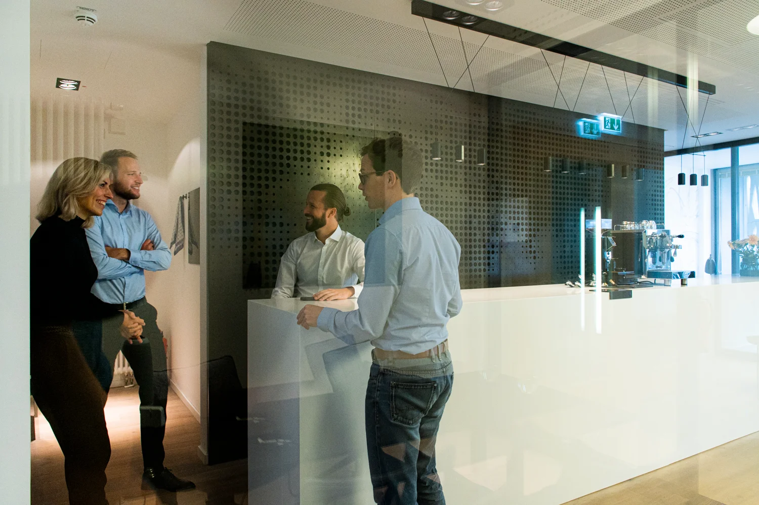 Team in conversation at a white counter, seen through a glass wall with reflections, Frankfurt am Main & Munich — Digital+, corporate photography by Janina Laszlo