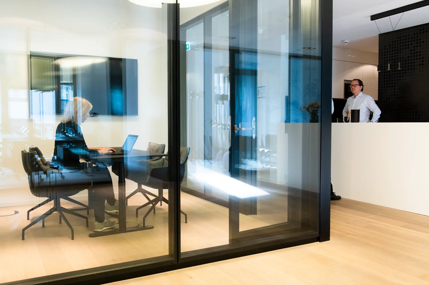Colleague working on a laptop inside a glass meeting room, a colleague standing at the counter behind, Frankfurt am Main & Munich — Digital+, corporate photography by Janina Laszlo