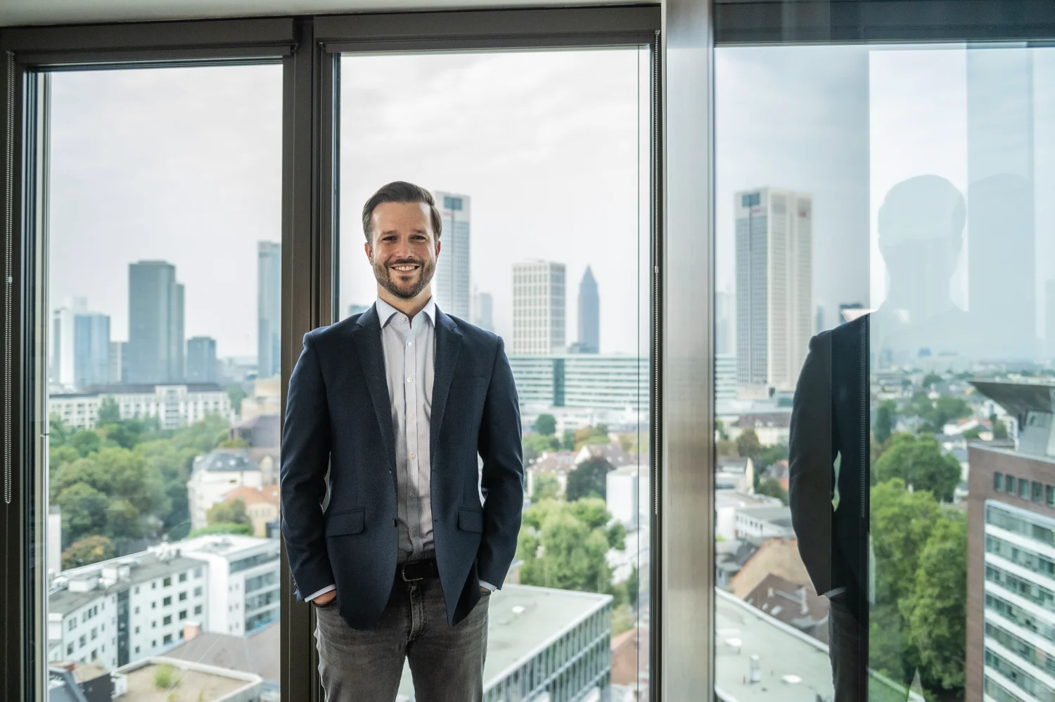 Employee in a navy blazer standing in front of a window with the Frankfurt skyline, business portrait, Frankfurt am Main — Digital+, corporate photography by Janina Laszlo